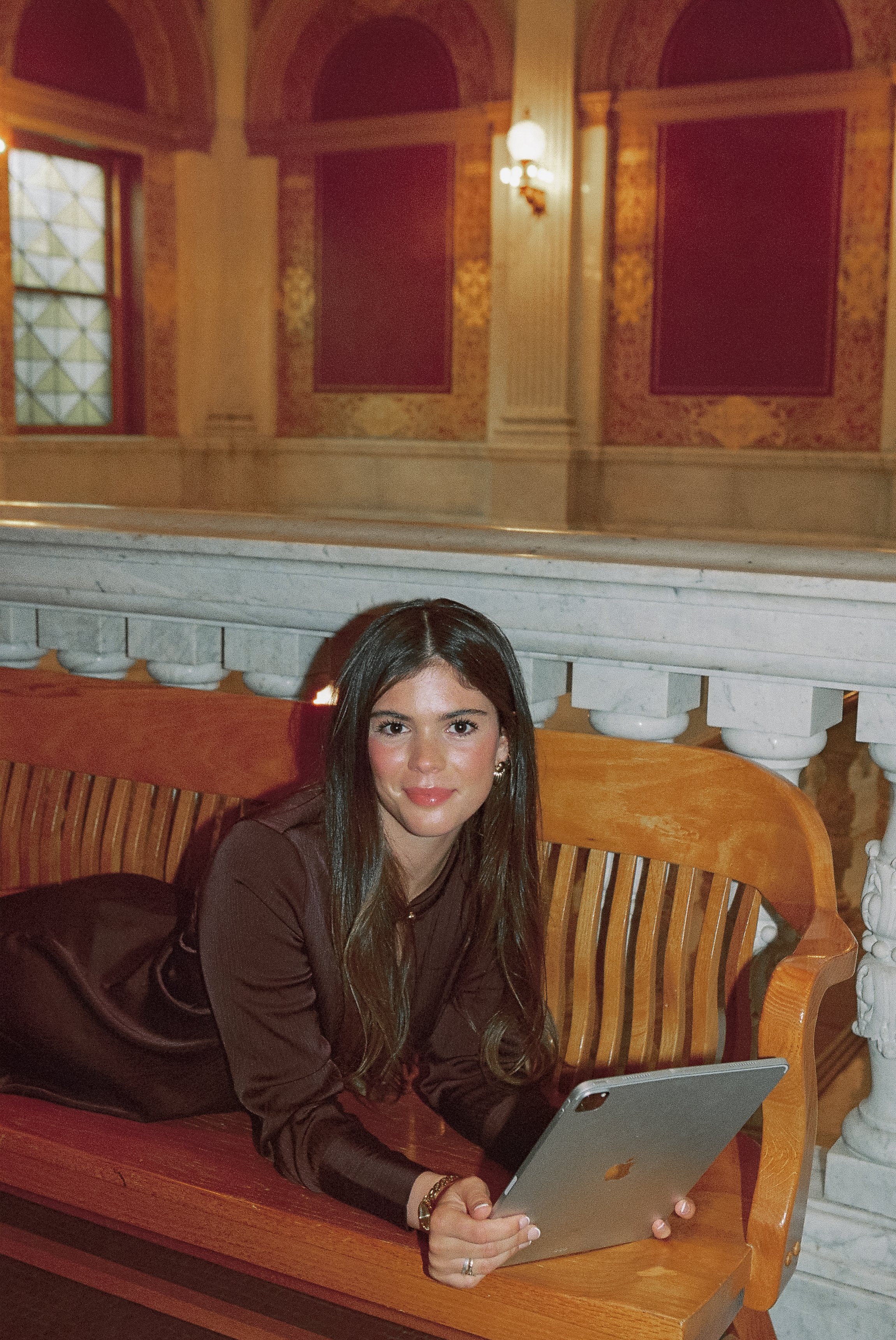 A woman with long dark hair and a brown blouse is sitting on a wooden bench holding a tablet, in an ornate room with gold-paneled walls and arched windows.