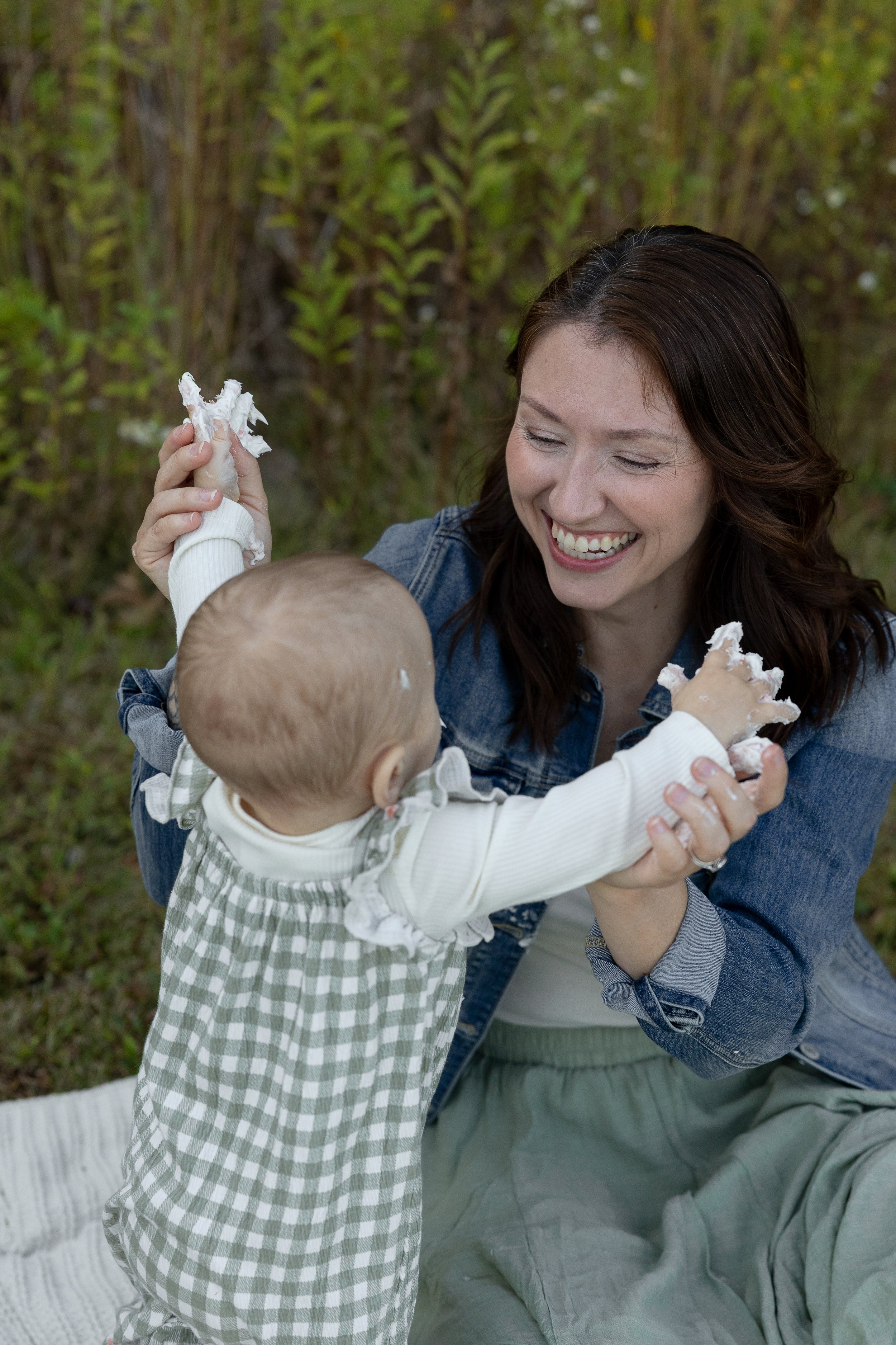 Young woman playing with a toddler, laughing, outdoors in a green, wooded area.
