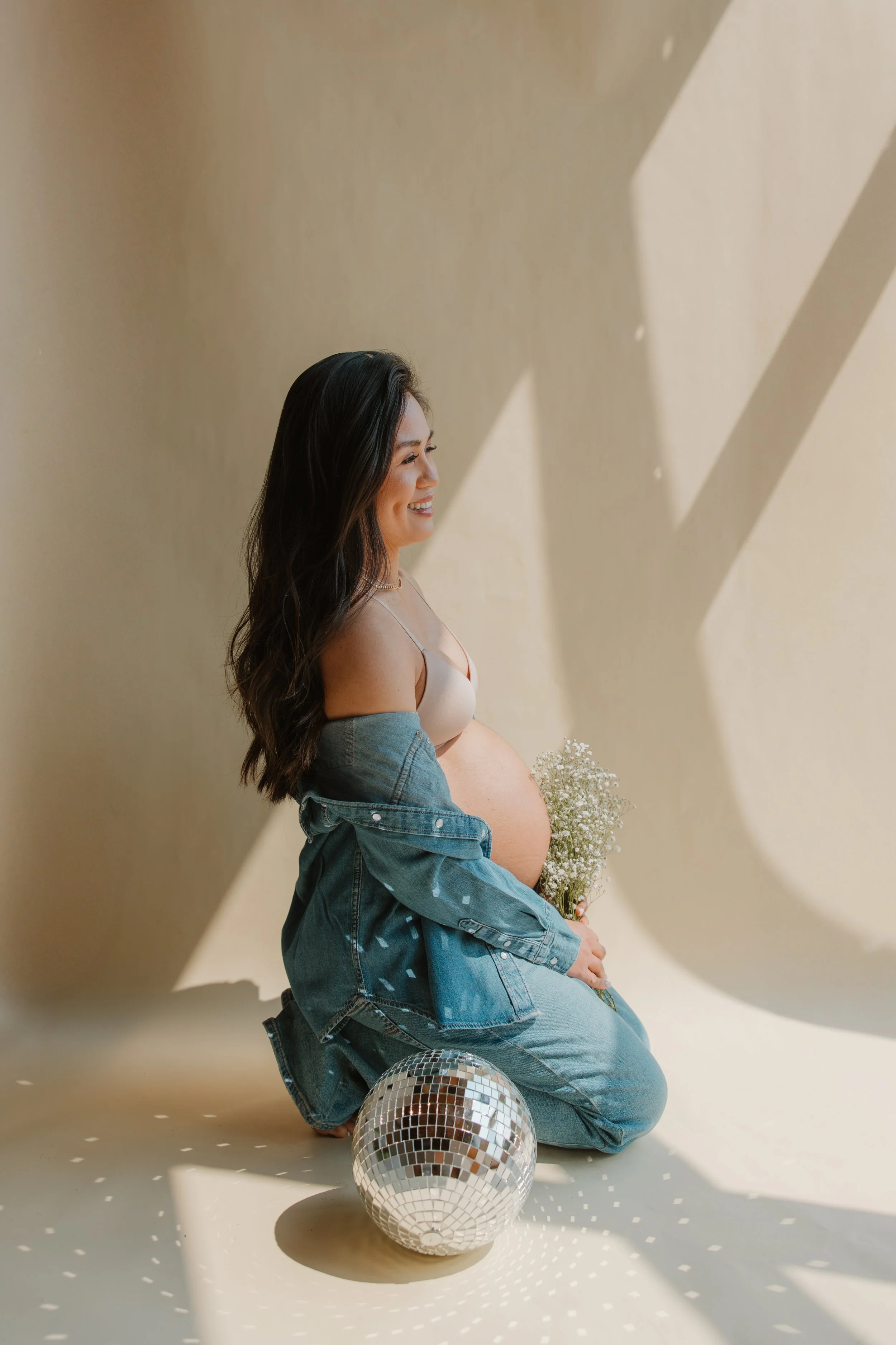 A pregnant woman with long dark hair, wearing a denim jacket off her shoulders and a white bra, smiling and holding a small bouquet of white flowers, sitting on the floor next to a disco ball, with sunlight creating geometric shadows on the beige wal