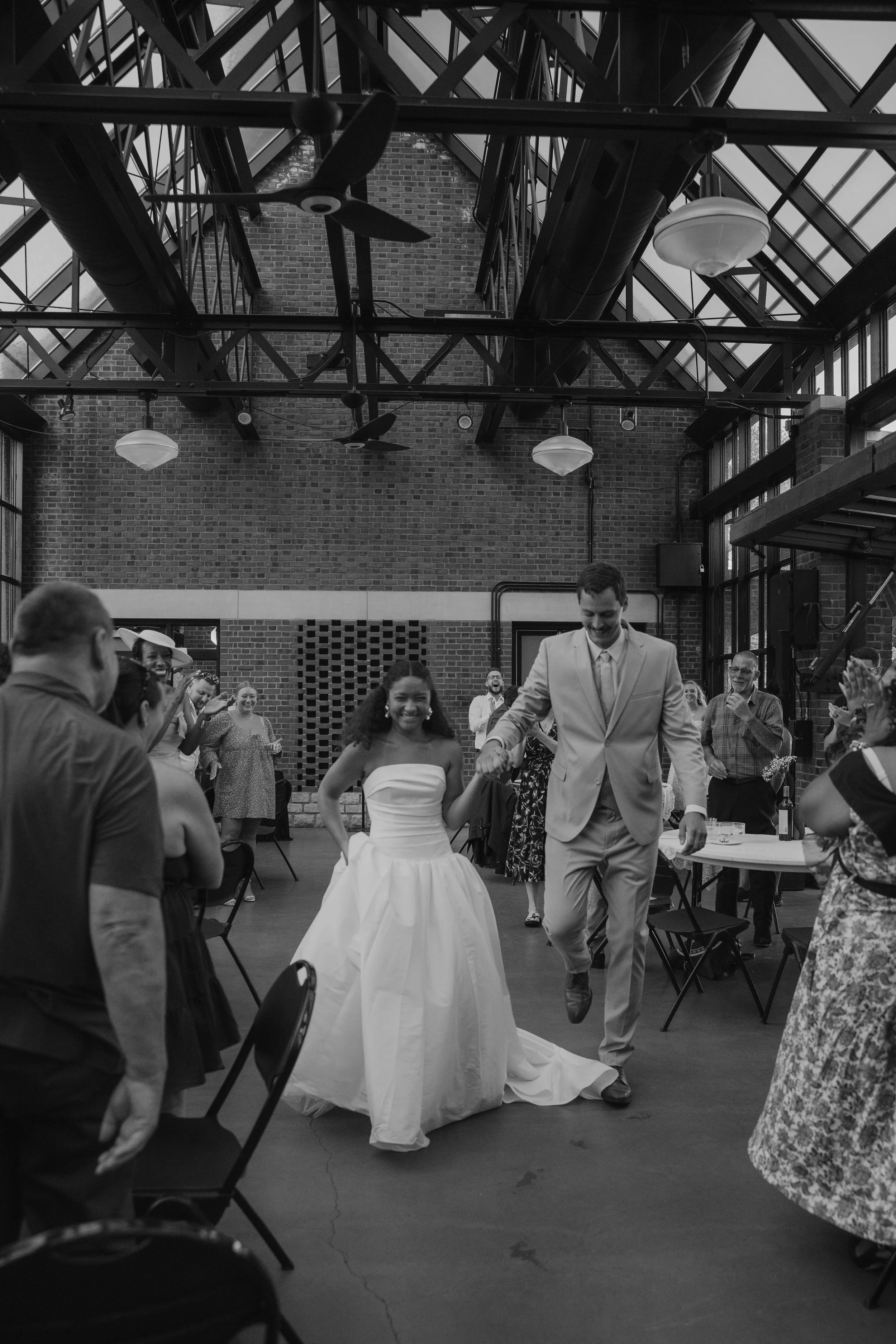 A bride and groom dancing at their wedding reception inside a venue with large windows and exposed brick walls, surrounded by their guests who are clapping and smiling.