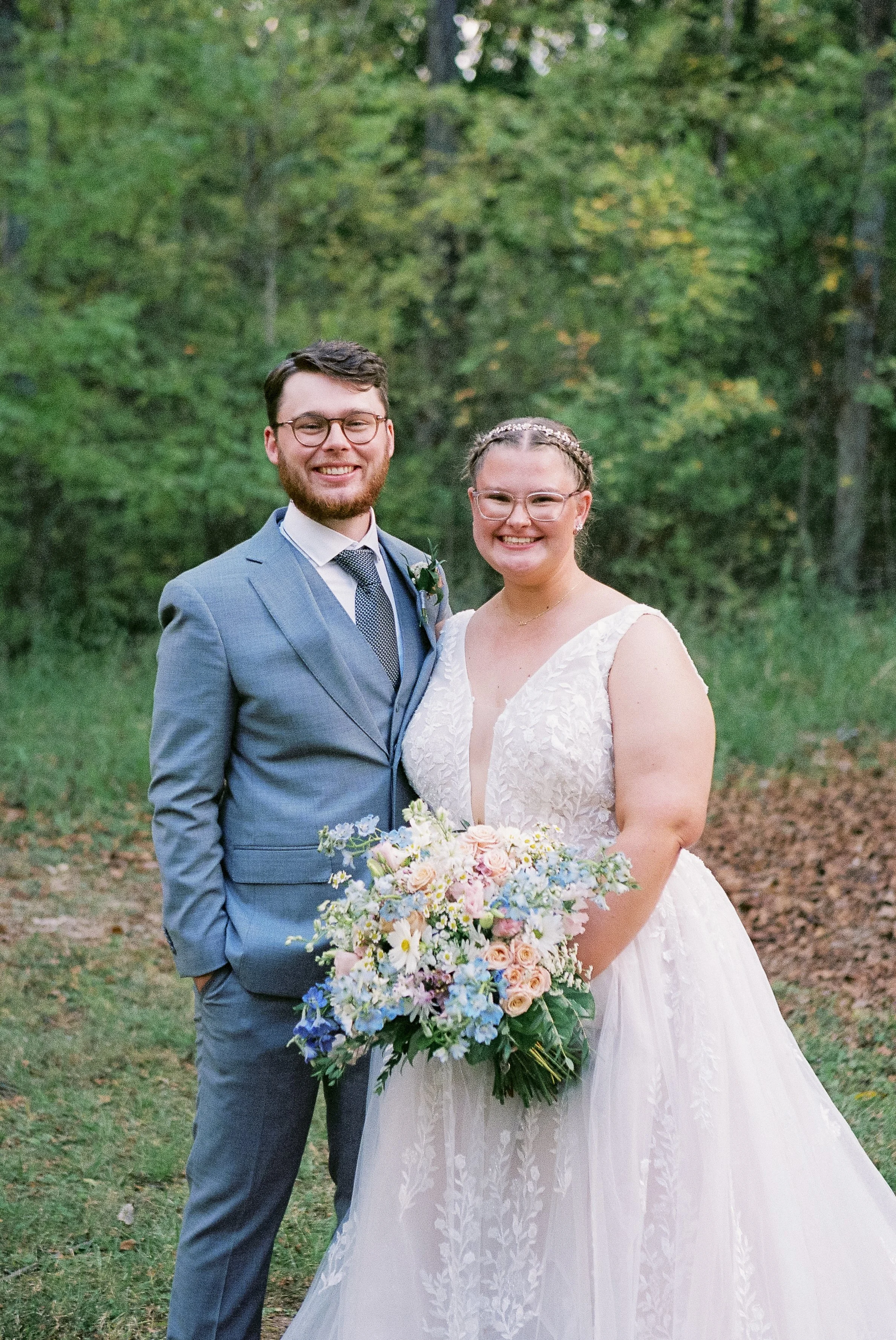 A newlywed couple standing outdoors surrounded by greenery. The groom has short dark hair, glasses, a beard, and is wearing a light blue suit with a white shirt and dark tie. The bride has her hair styled in braids, wears glasses, and a white wedding