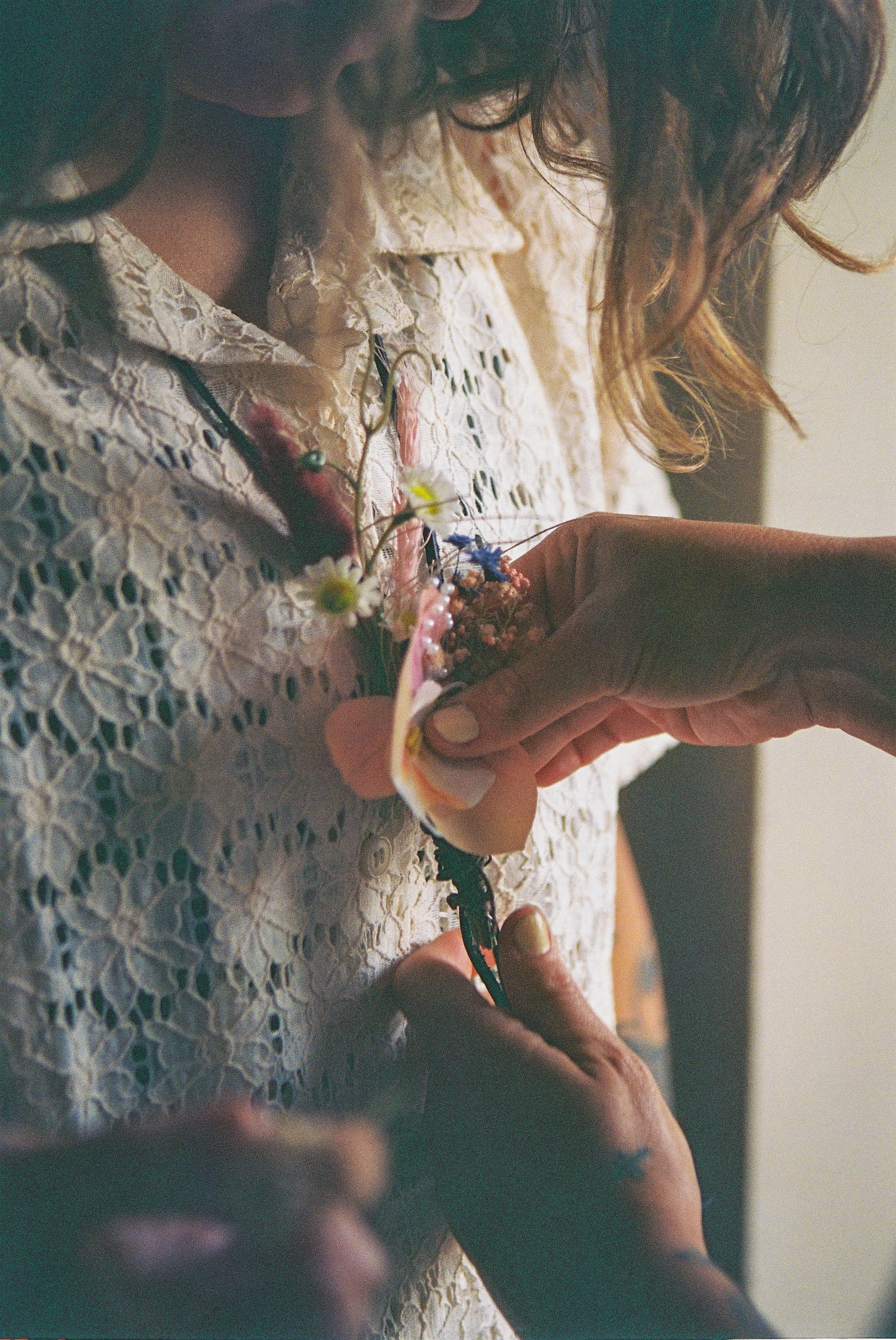 A person with curly hair fastens a boutonniere made of small flowers and greenery onto a lace dress.