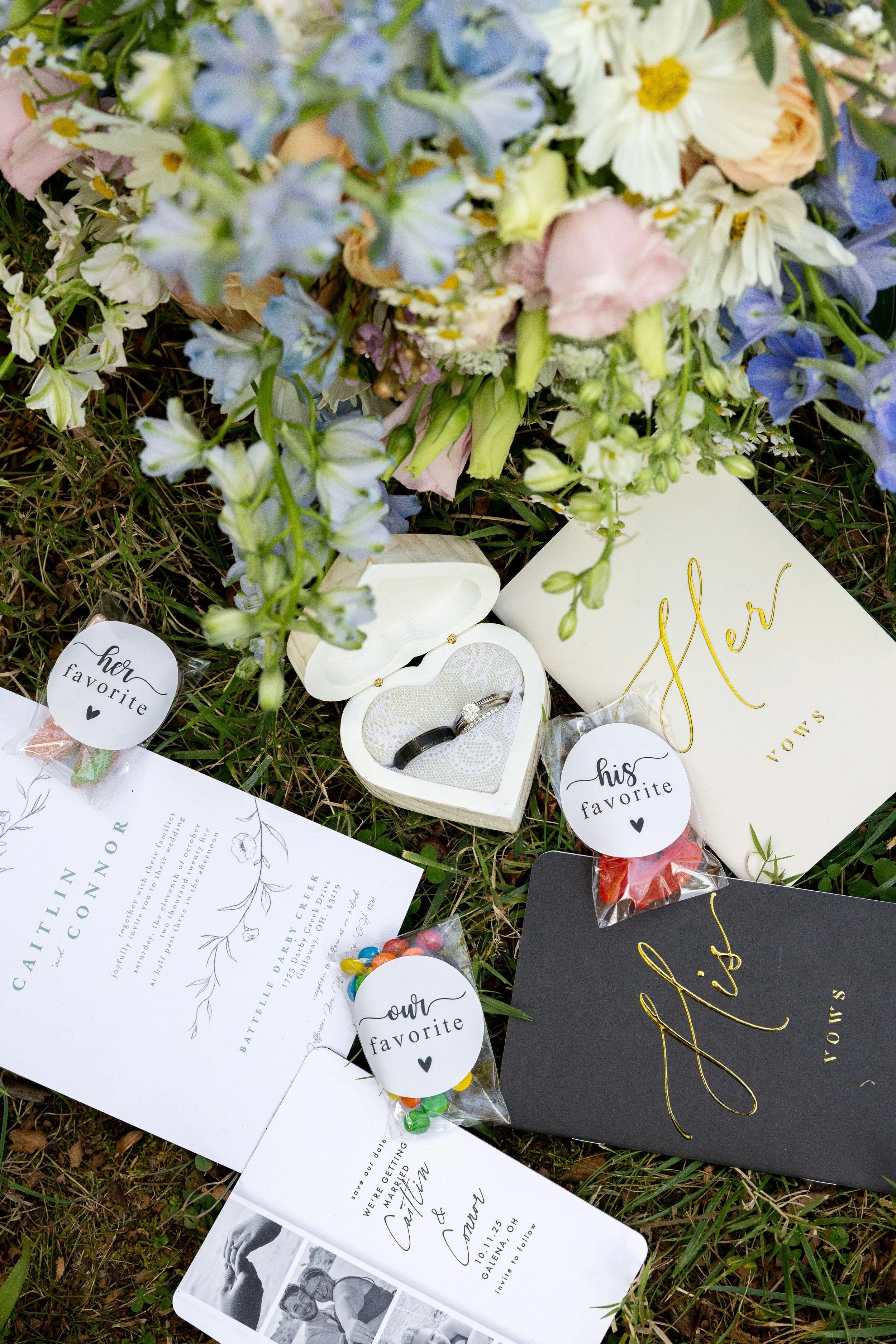 Wedding proposal items including a bouquet of mixed flowers, a white heart jewelry box with rings, and various cards and candies with labels 'her favorite,' 'his favorite,' and 'our favorite' placed on grass.