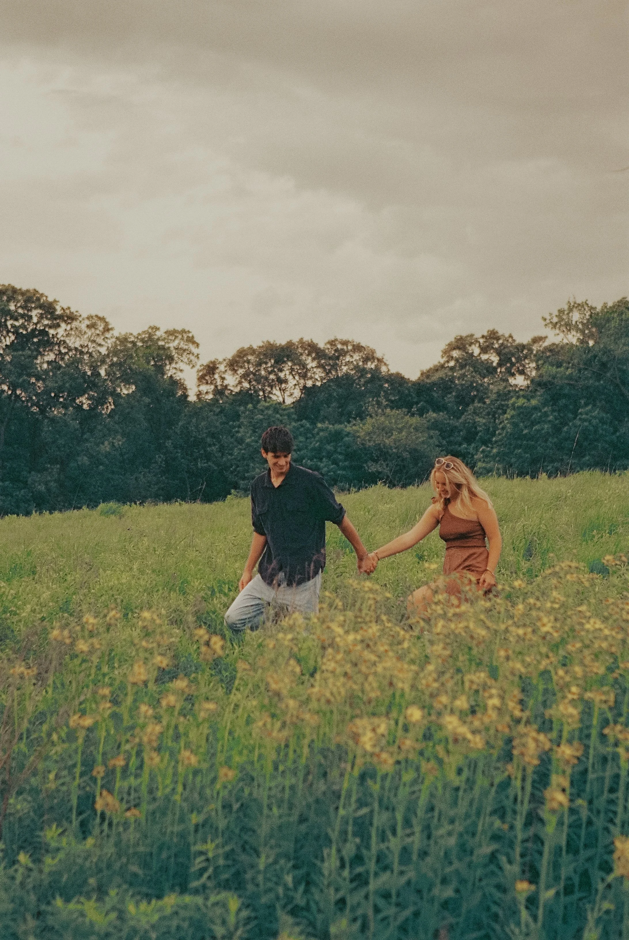 A man and a woman holding hands and walking through a grassy field with trees in the background.