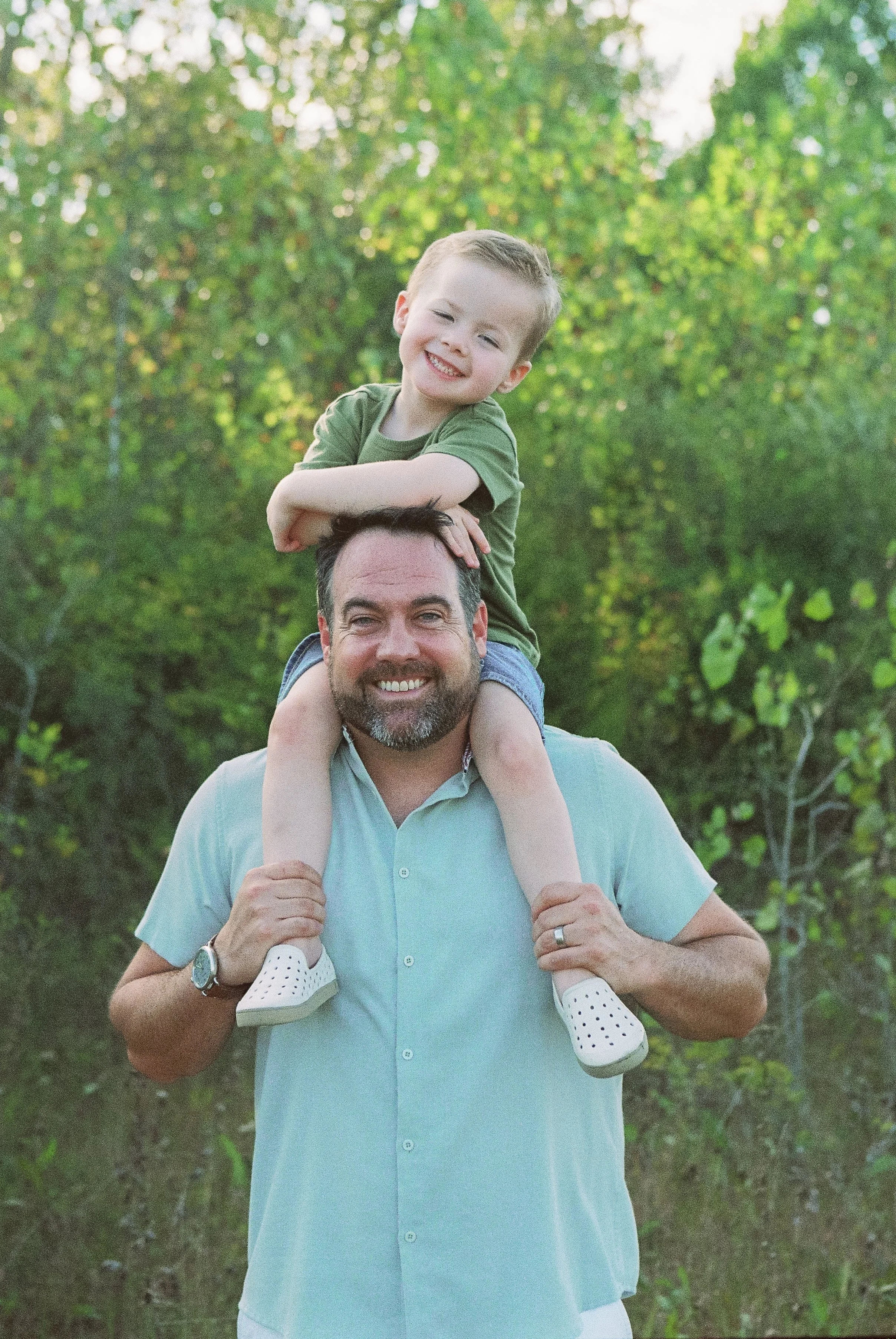 A man carrying a young boy on his shoulders in a wooded outdoor setting, both smiling.