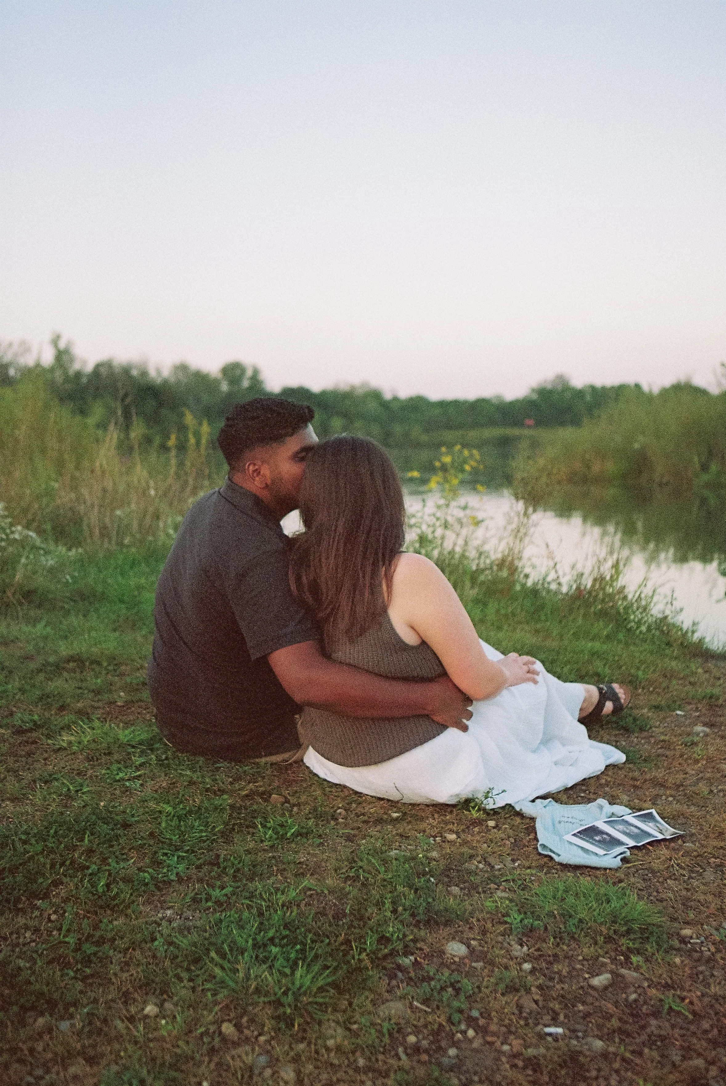 A couple sitting by a river during sunset, sharing a moment and looking into the distance.