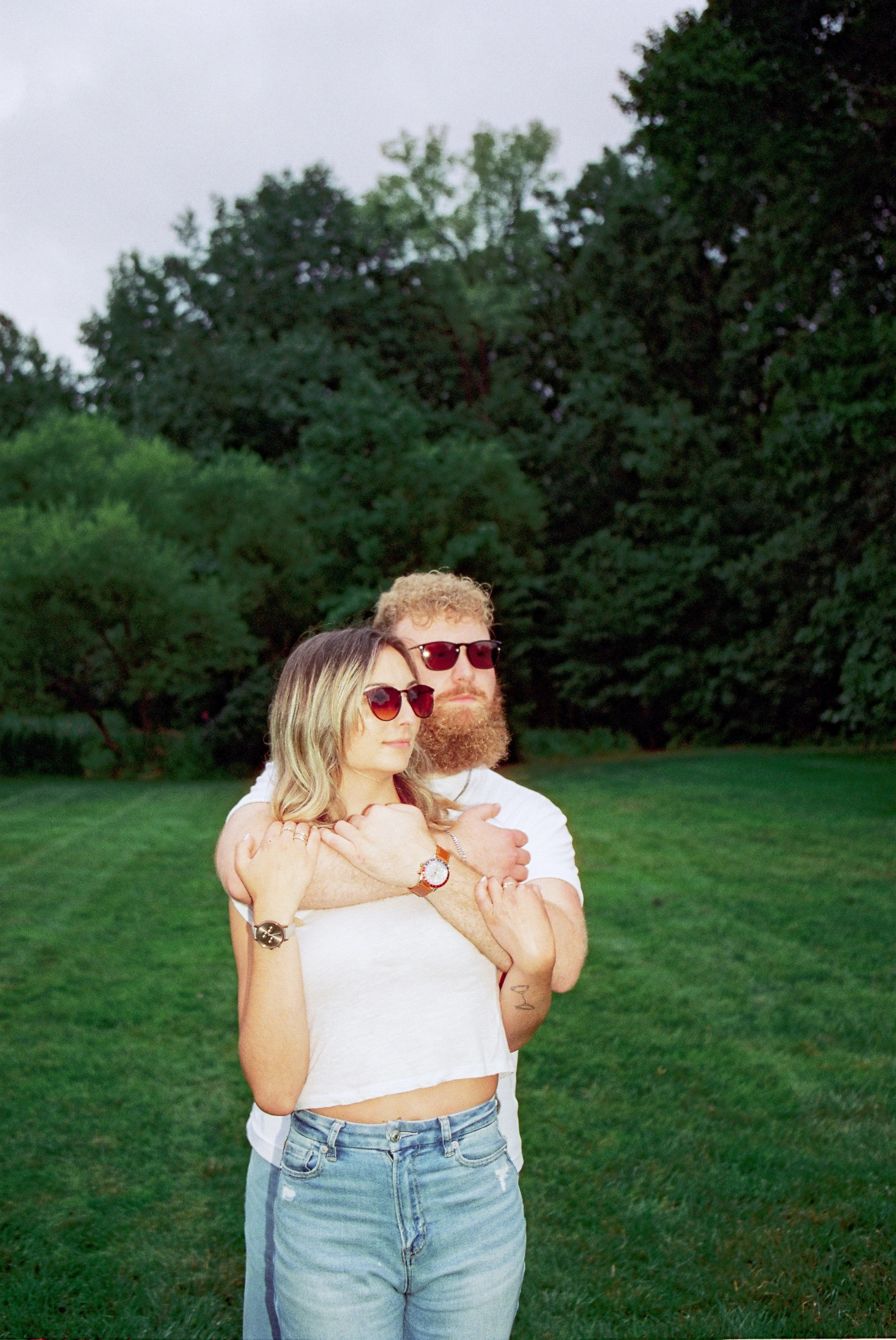 A man with curly hair and a beard and a woman with blonde hair and sunglasses standing outdoors on a grassy area surrounded by trees, with the man embracing the woman from behind.