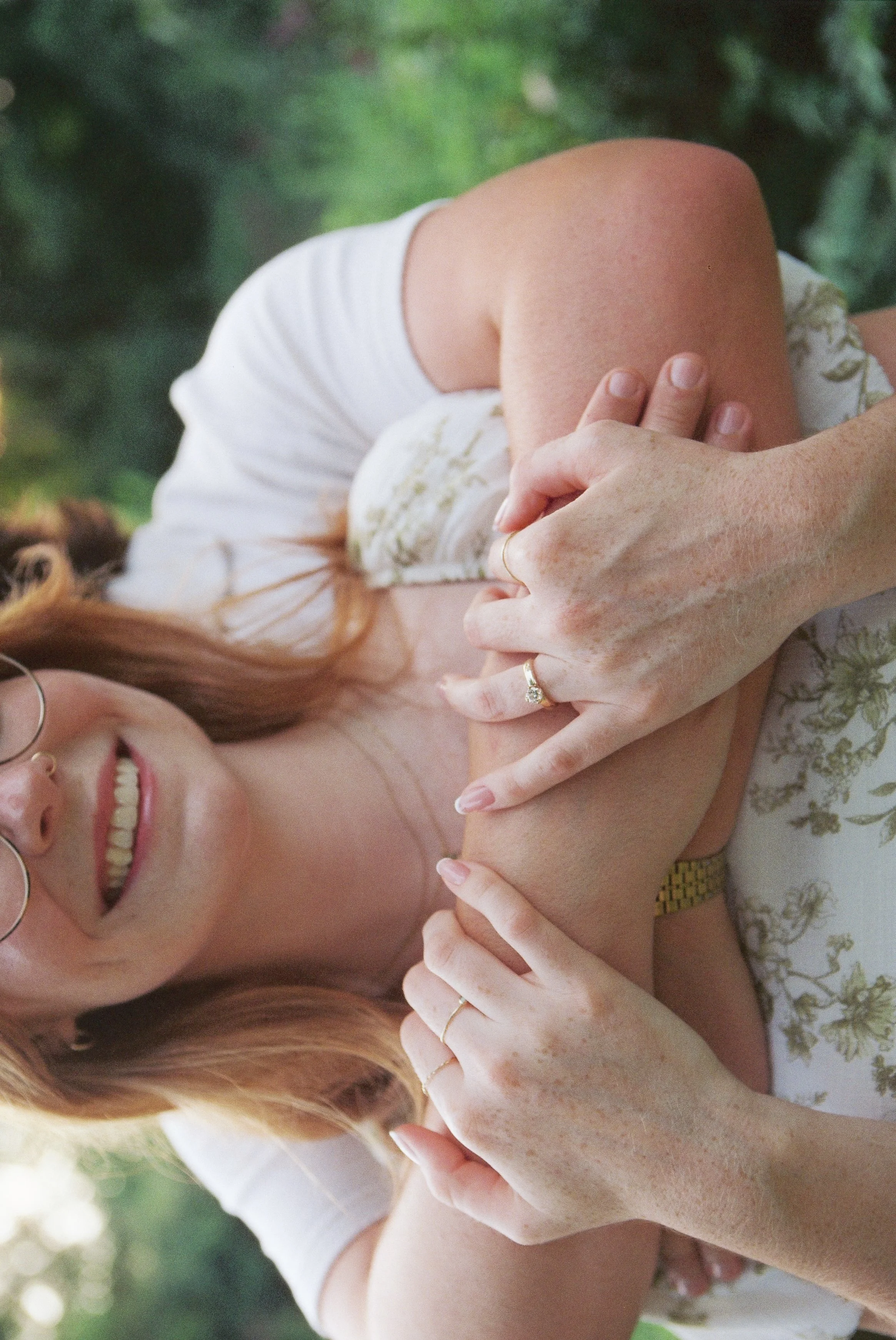 A woman with red hair, glasses, and freckles is smiling and lying down outdoors. She is wearing a white dress with a yellow floral pattern and some jewelry, including rings and a watch. Multiple hands are touching her arm and shoulder, showing affect