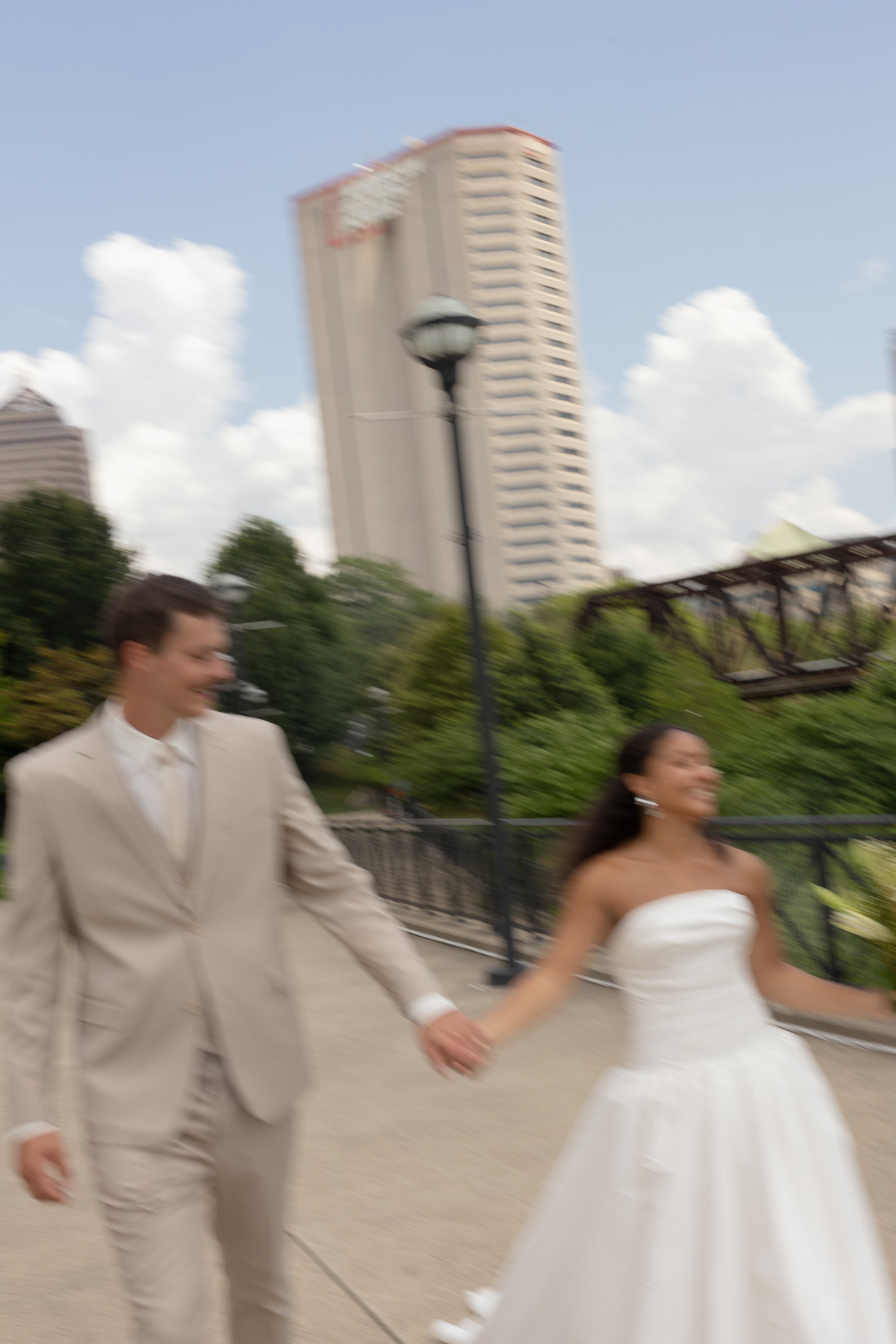 Blurred photo of a couple holding hands and walking outdoors, with a tall building in the background. The woman is wearing a white strapless dress, and the man is in a beige suit.