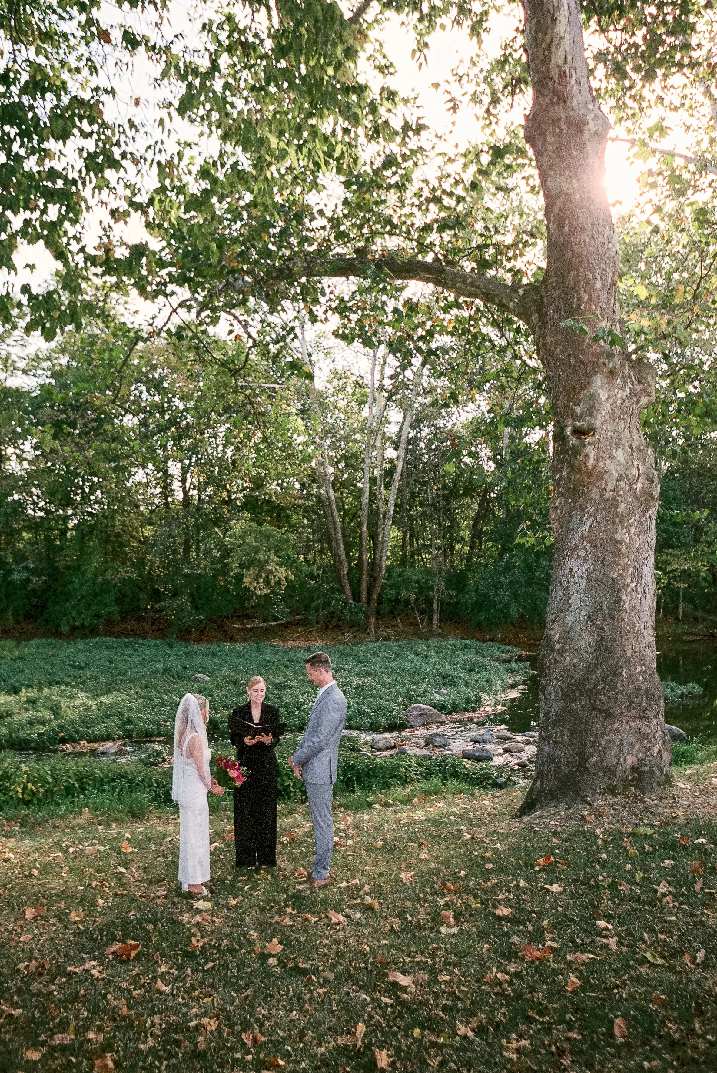 A wedding ceremony taking place outdoors by a river at sunset with a bride in a white dress and veil, a groom in a gray suit, and an officiant dressed in black, all standing on grass surrounded by trees.