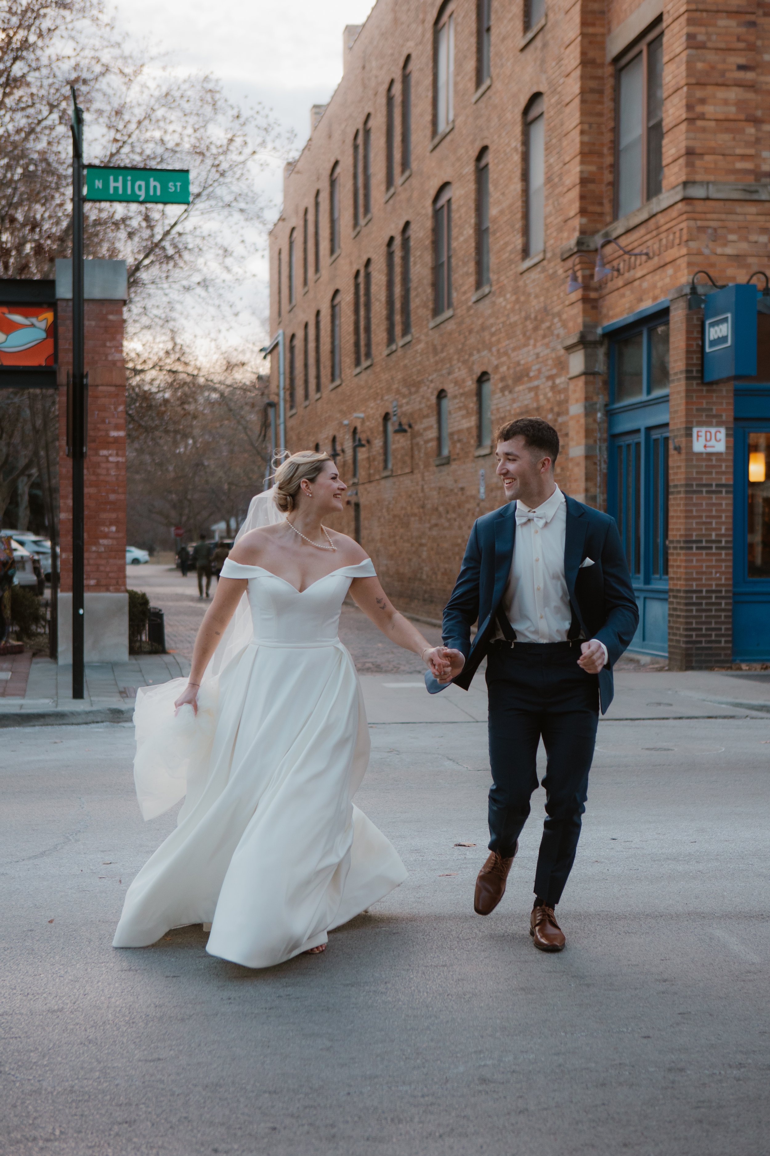 A bride and groom in wedding attire holding hands and running on a city street, smiling at each other.