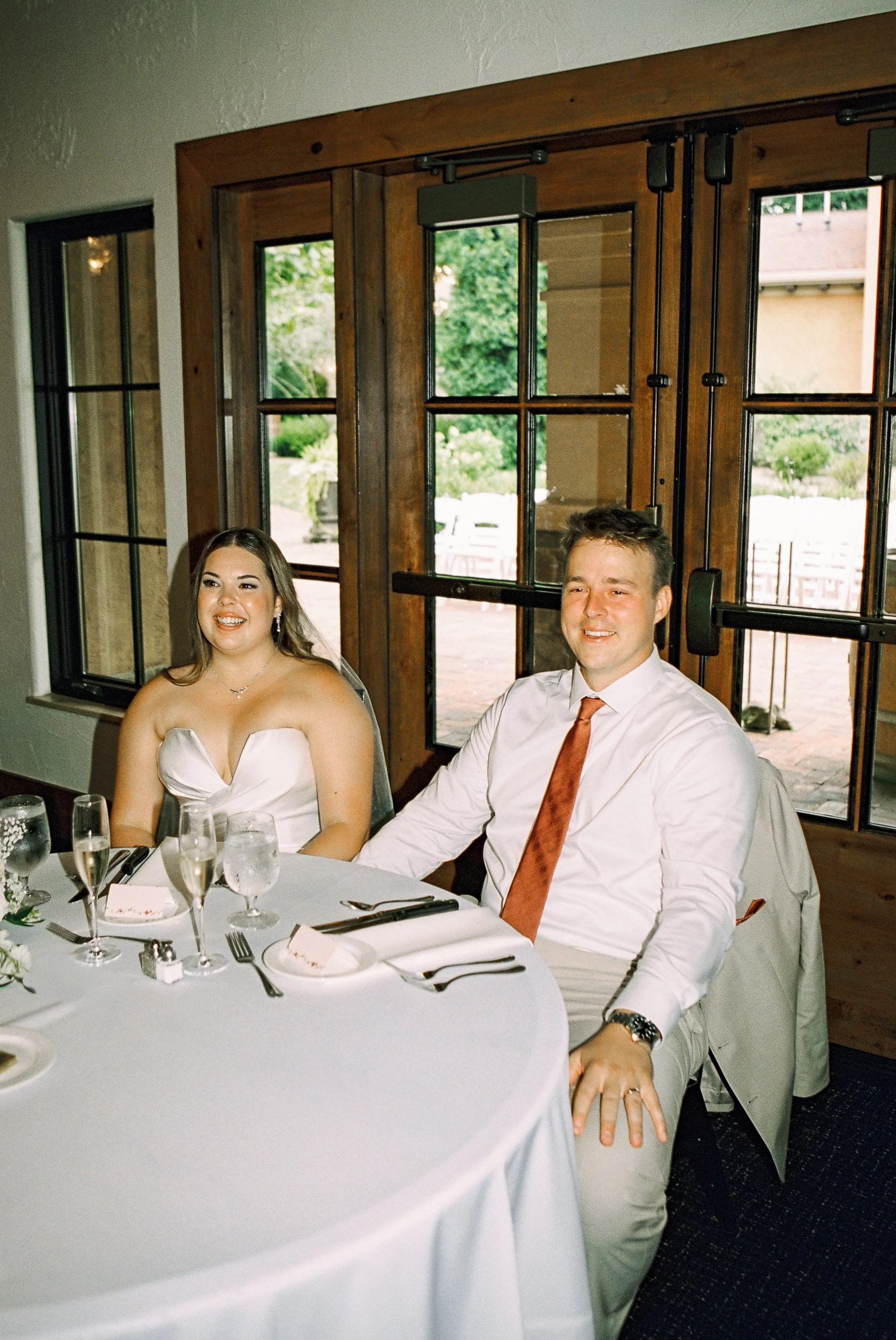 A smiling woman in a strapless white dress and a smiling man in a white shirt with an orange tie sitting at a round table during a celebration.