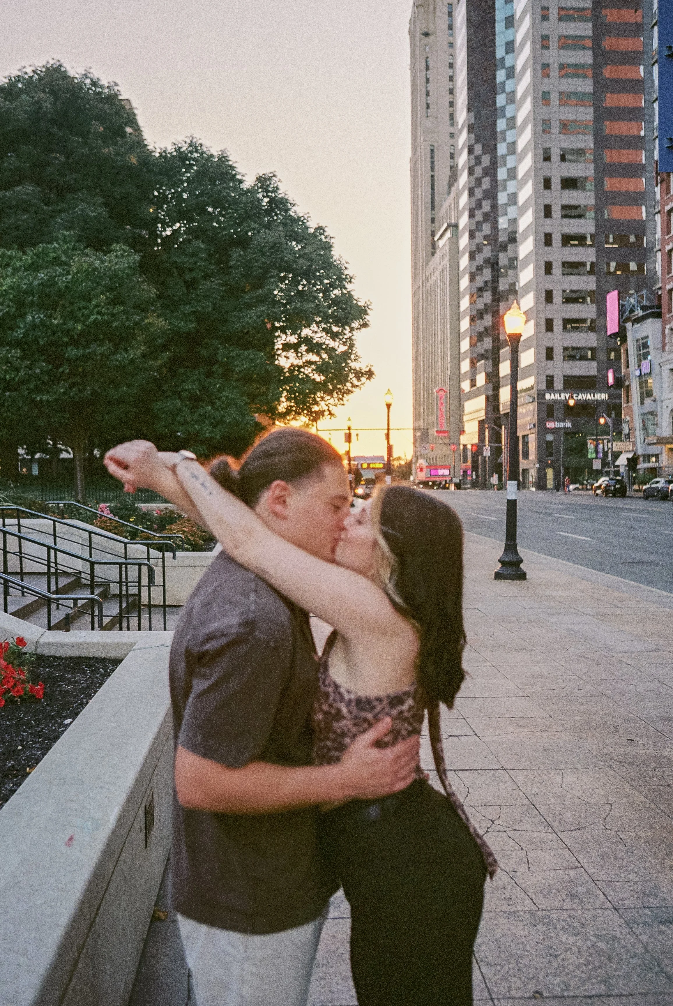 A couple sharing a kiss on a city street during sunset, with tall buildings and streetlights in the background.