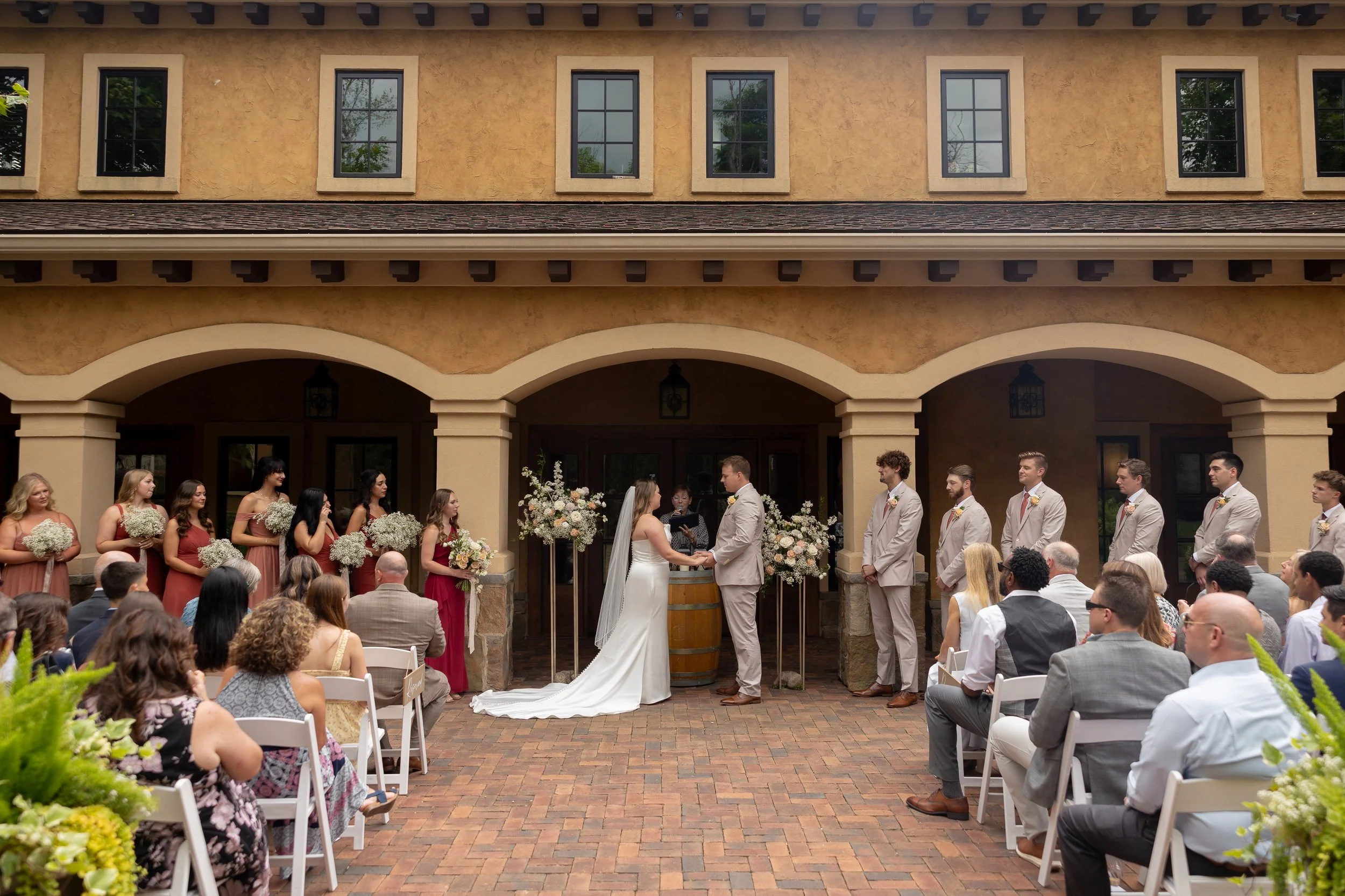 A wedding ceremony outdoors with the bride and groom holding hands in front of an officiant, surrounded by bridesmaids in burgundy dresses and groomsmen in beige suits, all under an archway in front of a tan building with multiple windows, with seate