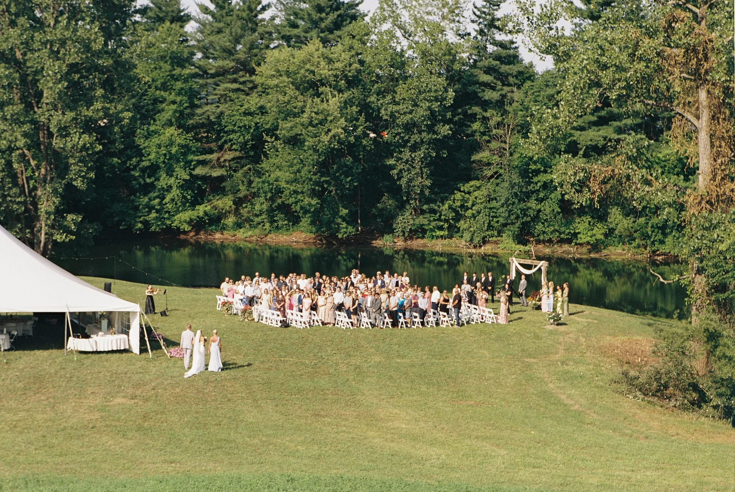Outdoor wedding ceremony on a grassy area near a lake, with guests seated in white chairs under a wooden wedding arch, surrounded by green trees.