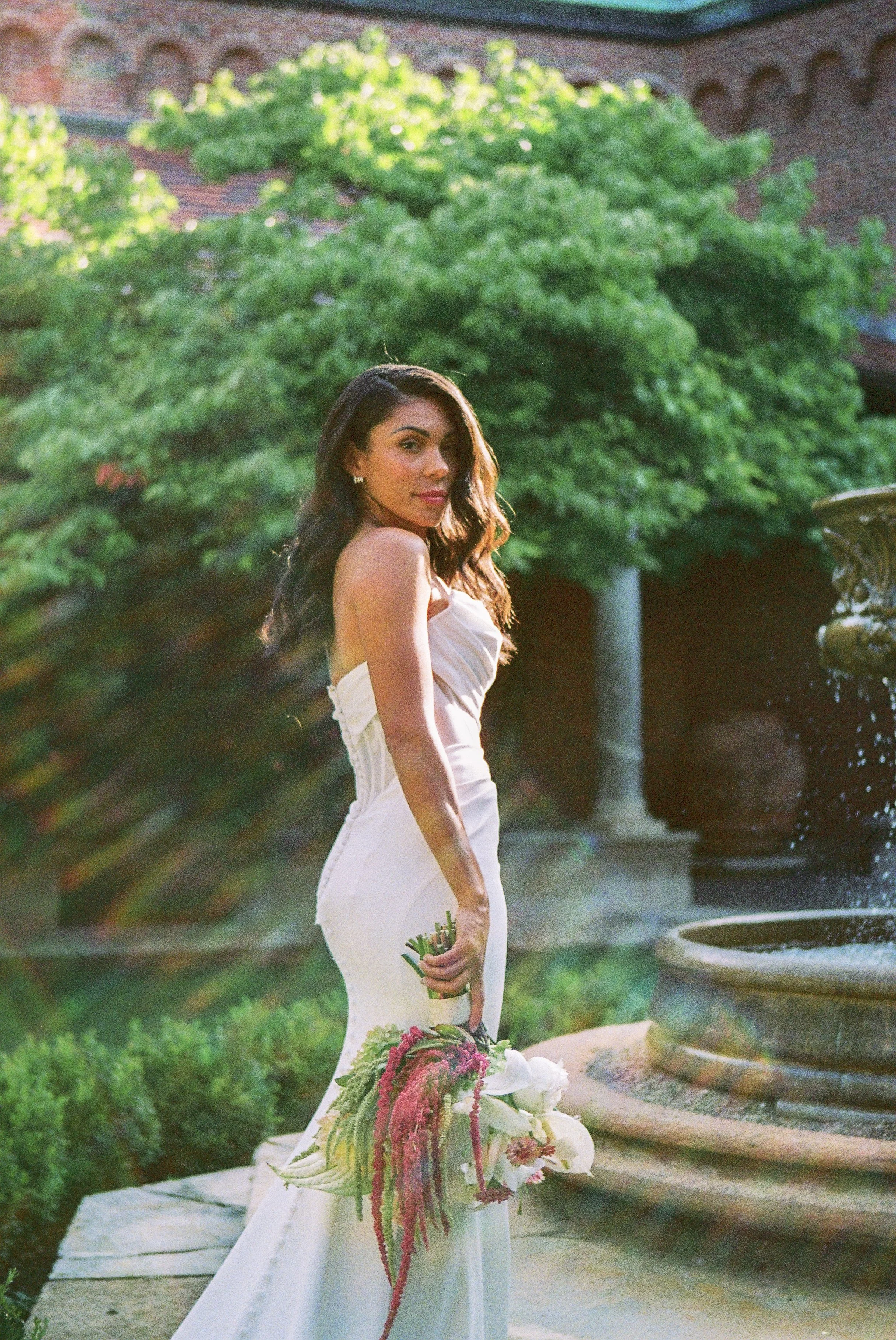 A woman in a strapless white dress holding a bouquet of colorful flowers, standing near a water fountain in a garden with lush green trees.
