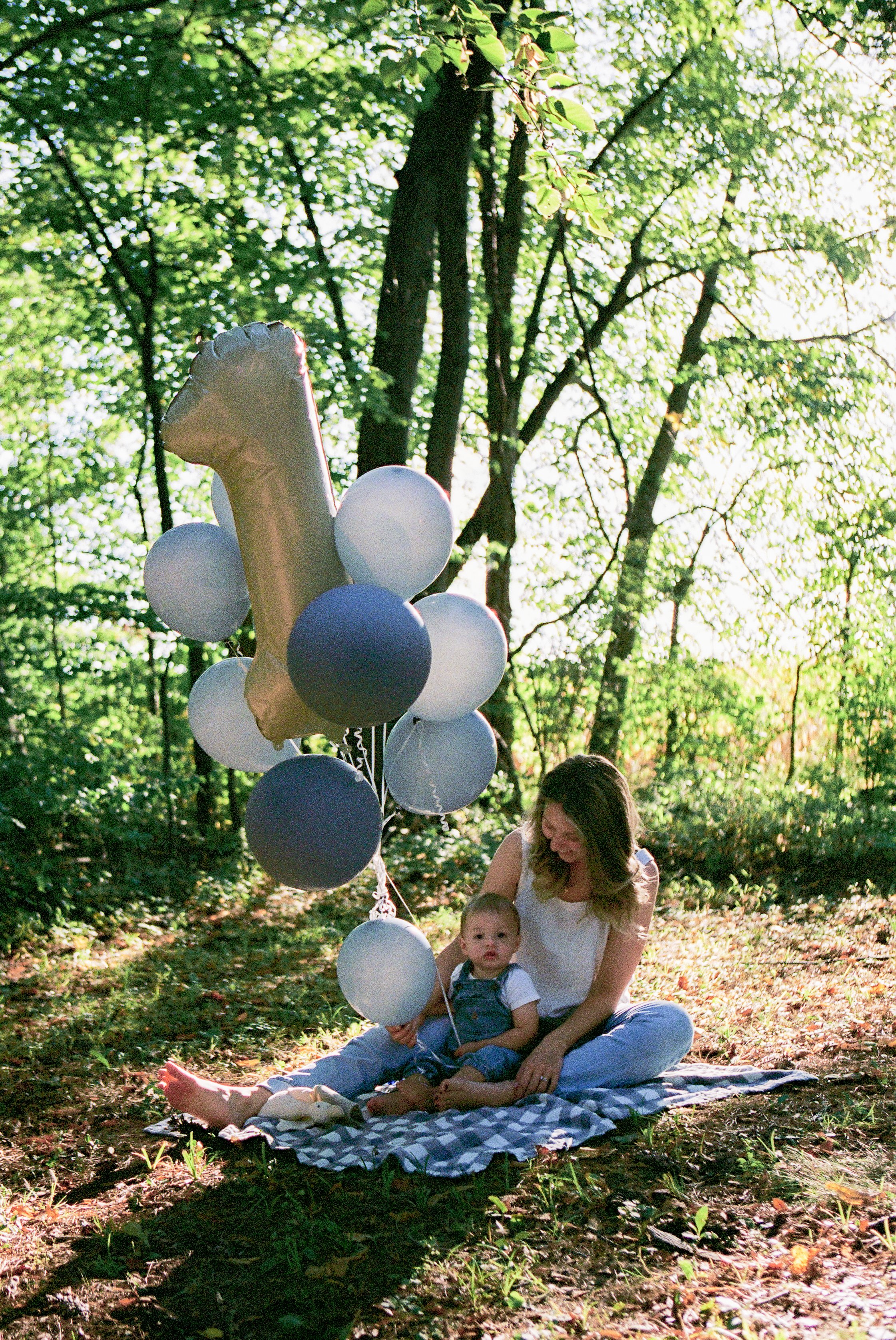 A woman and a young child sitting on a checkered blanket in a sunlit forest, holding a bunch of balloons, including a large balloon shaped like a number one.