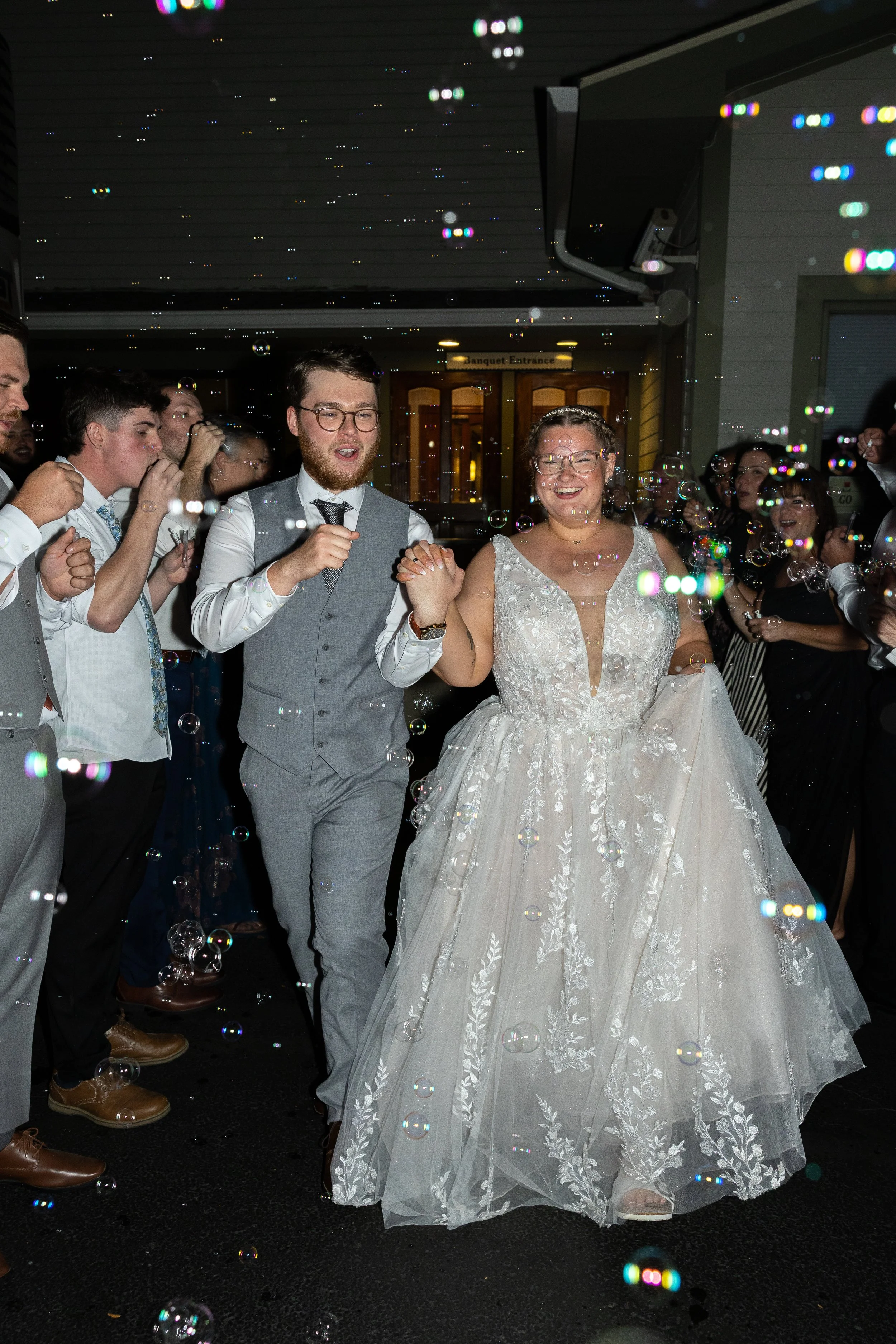 Bride and groom holding hands, walking through bubbles at their wedding reception. Guests are standing around, some blowing bubbles or taking photos, indoor venue with a wooden door in the background.