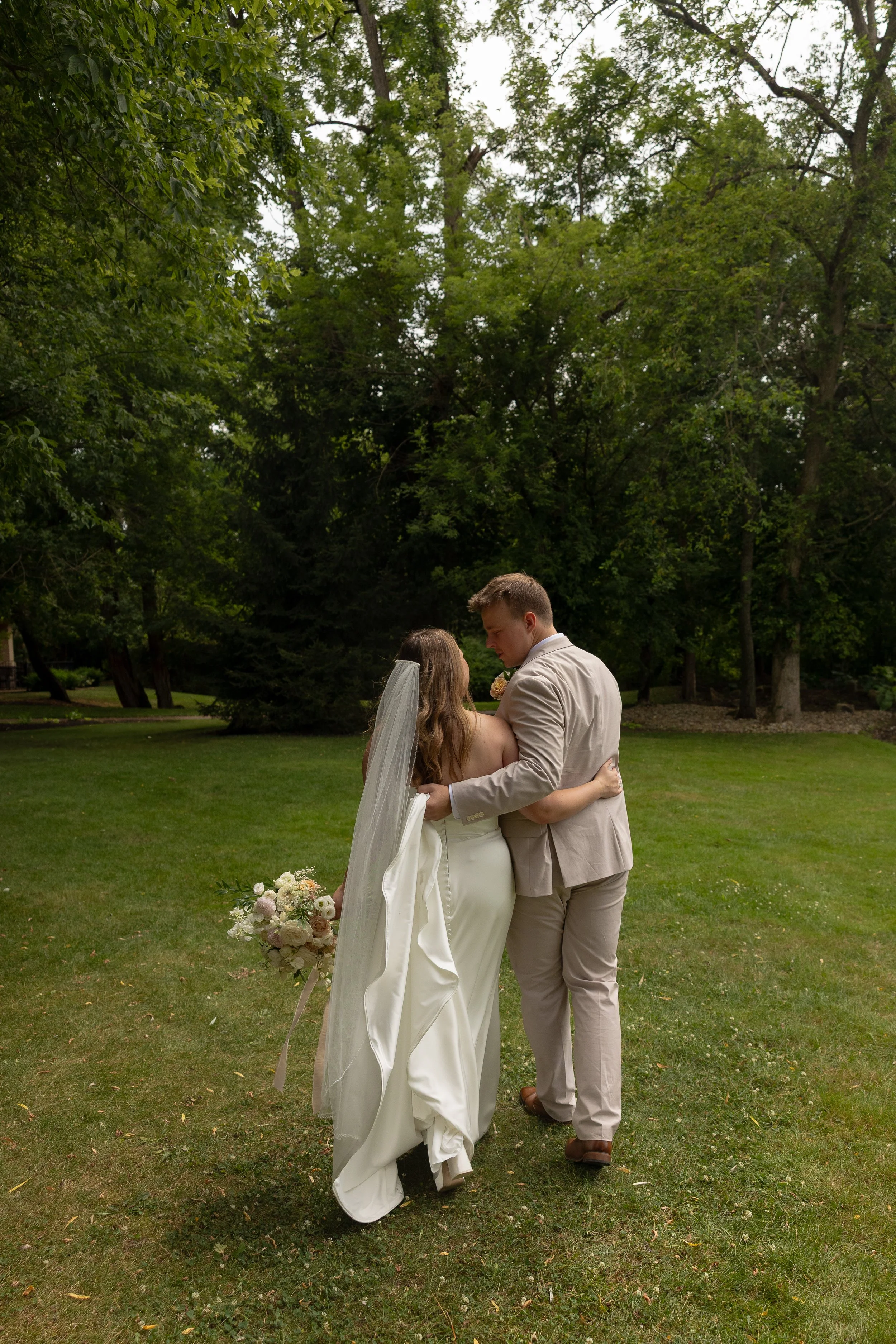 A bride and groom embrace outdoors in a lush green park, with the bride in a white wedding dress and veil holding a bouquet, and the groom in a light-colored suit.