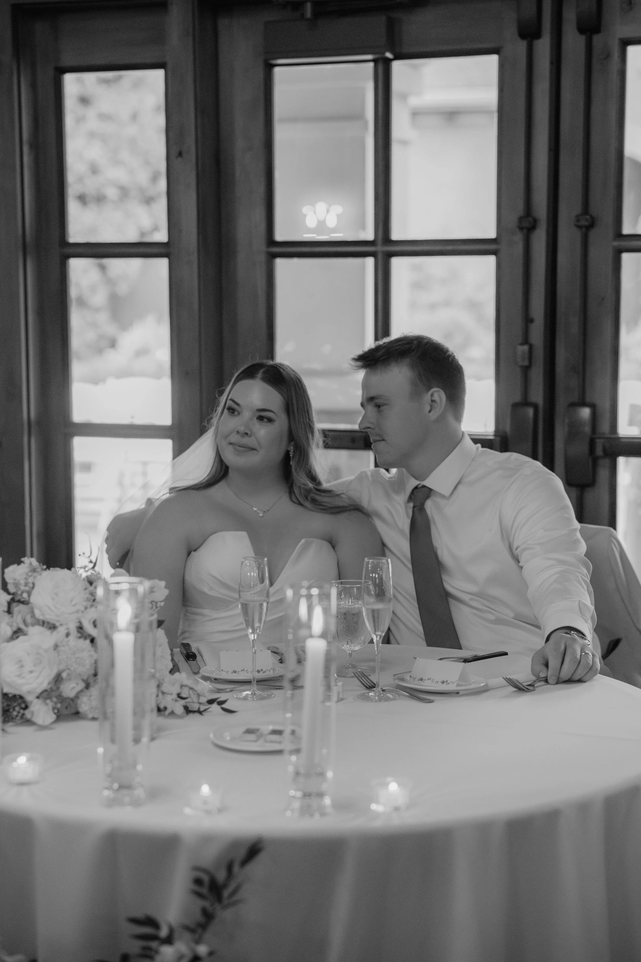 Black and white photo of a bride and groom sitting at a decorated wedding reception table, with candles and floral arrangements, indoors near large windows.
