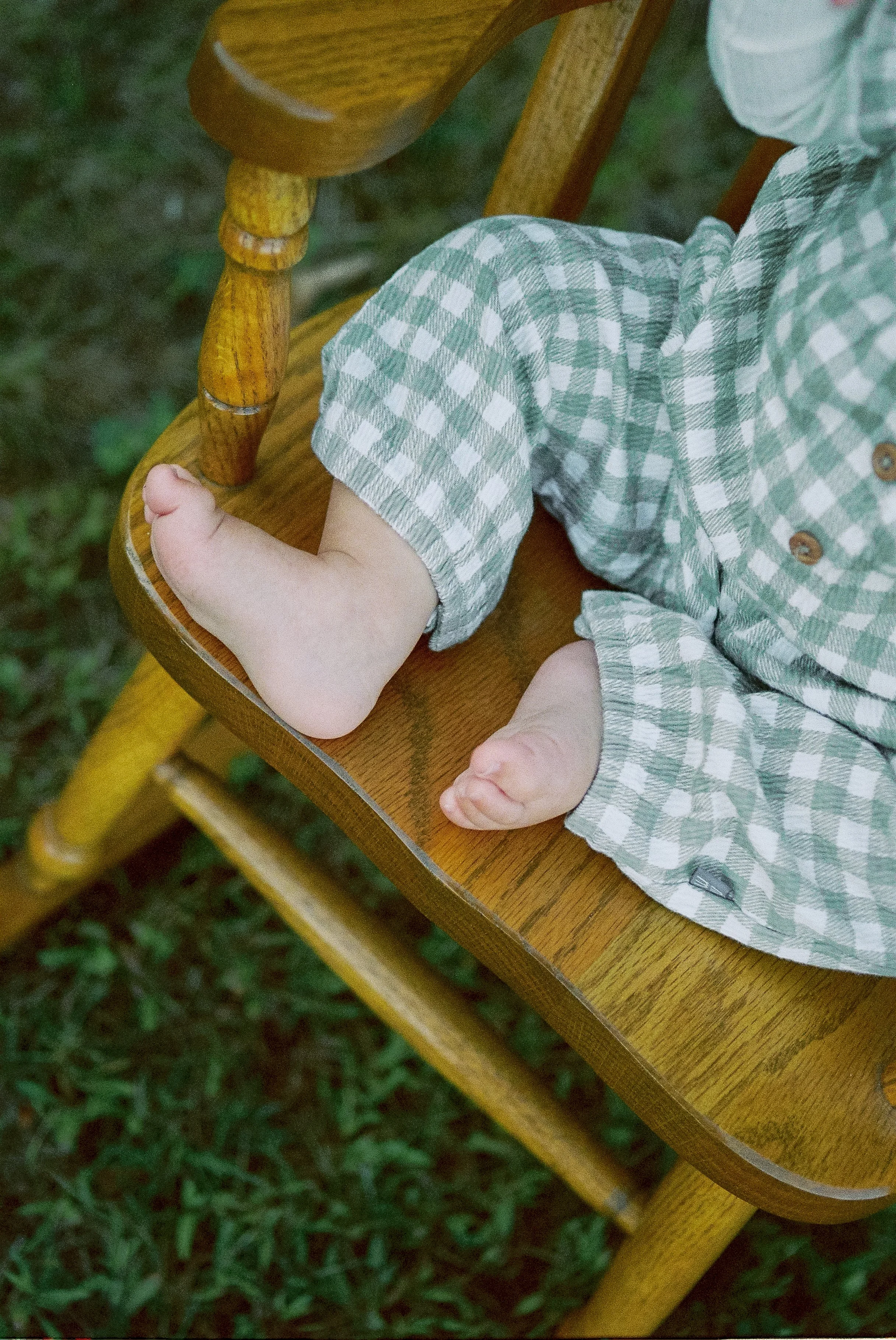 Close-up of a baby sitting in a wooden high chair, wearing a green and white checkered outfit, with bare feet resting on the seat.