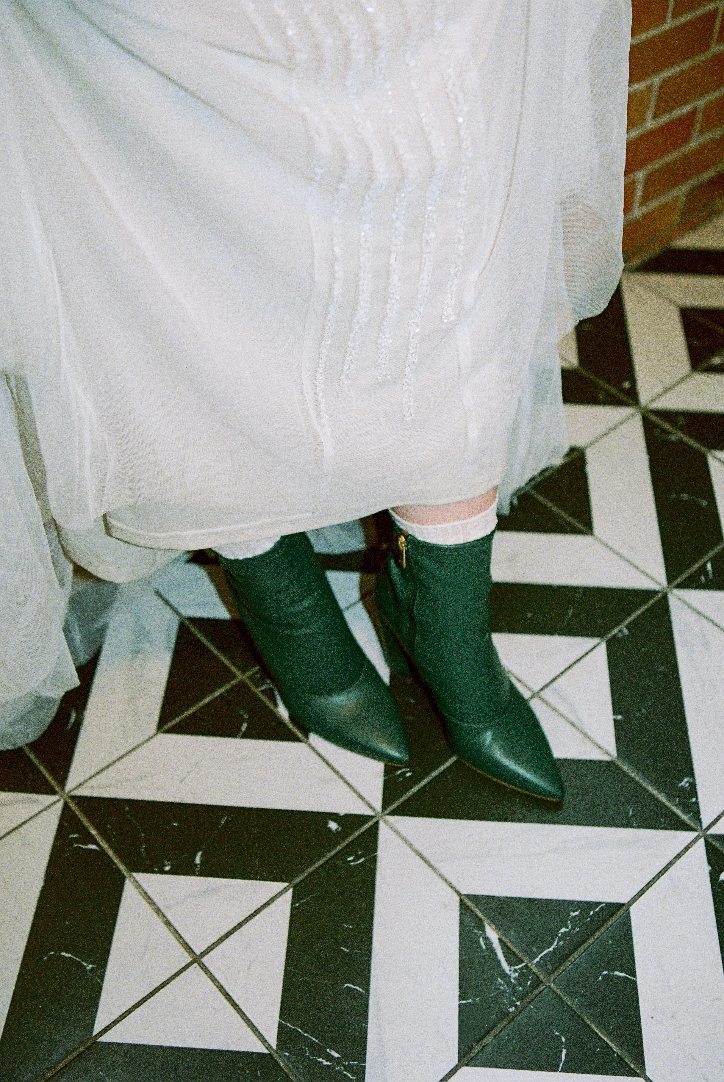 A woman wearing a white dress with embroidery, knee-high socks, and black high-heeled boots standing on a black and white geometric tiled floor.