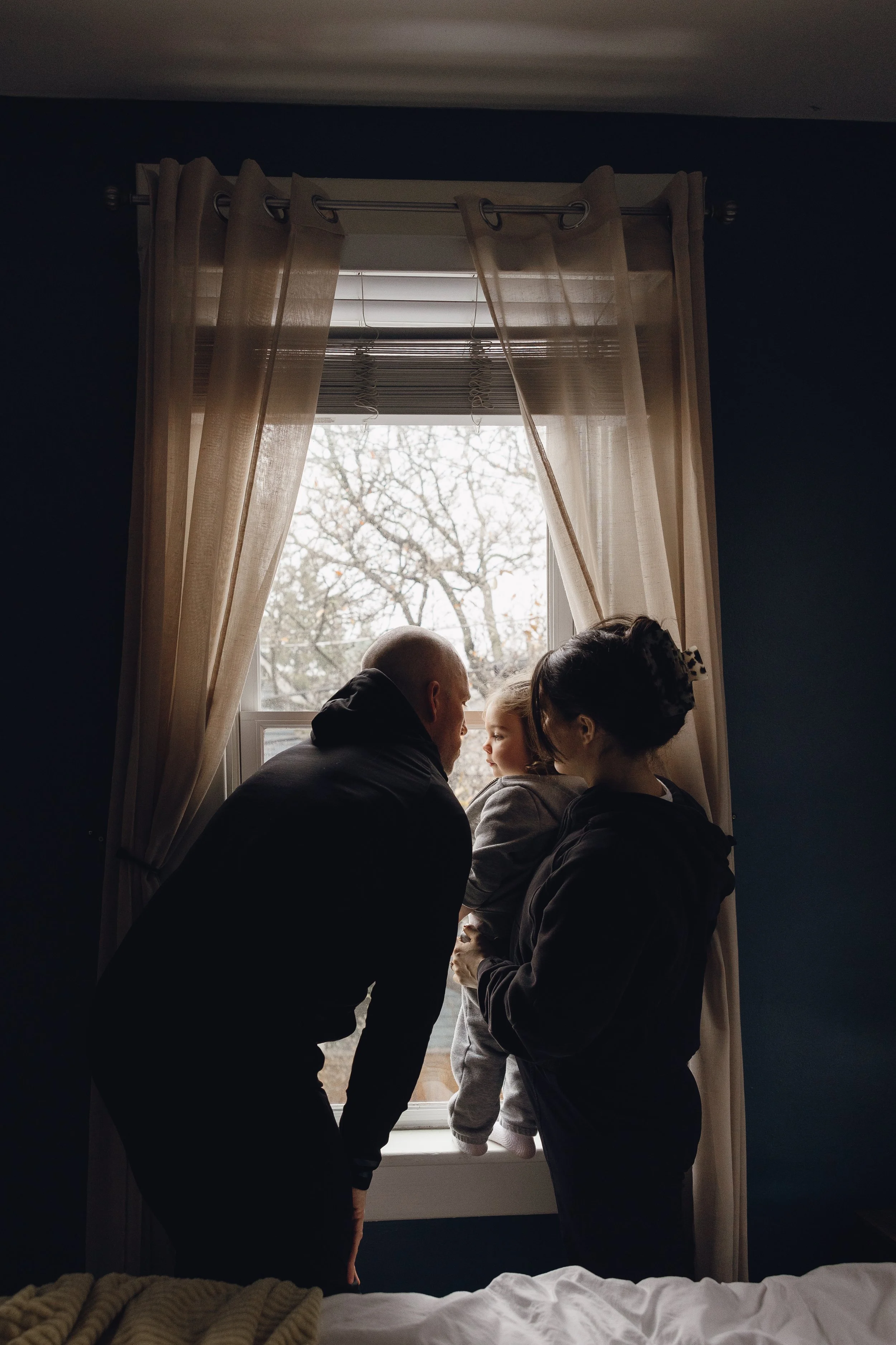 A family of three looking out of a window in a dimly lit room, with trees visible outside.