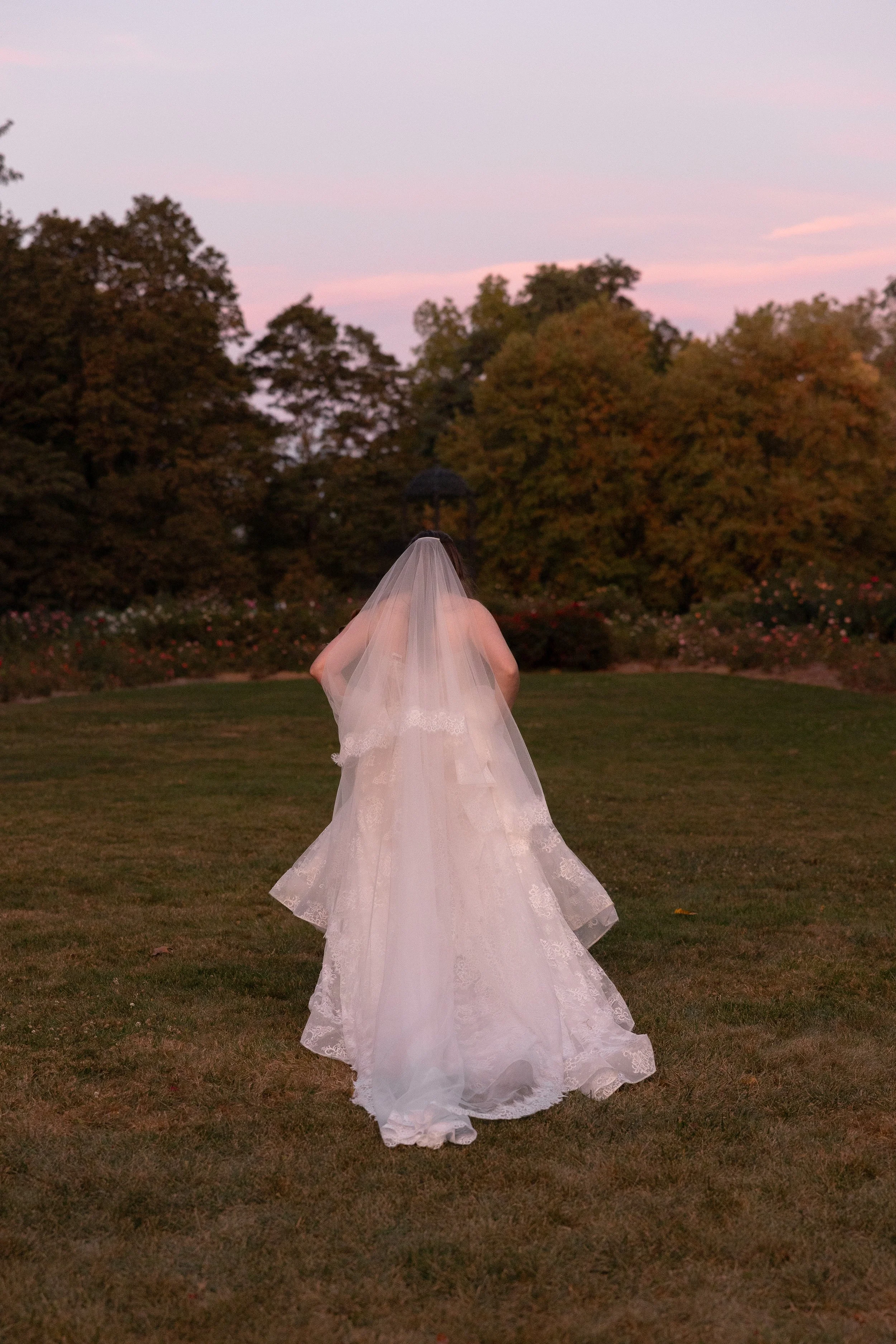 Bride in a wedding gown with a veil walking on grass at sunset in a park with trees and pink sky