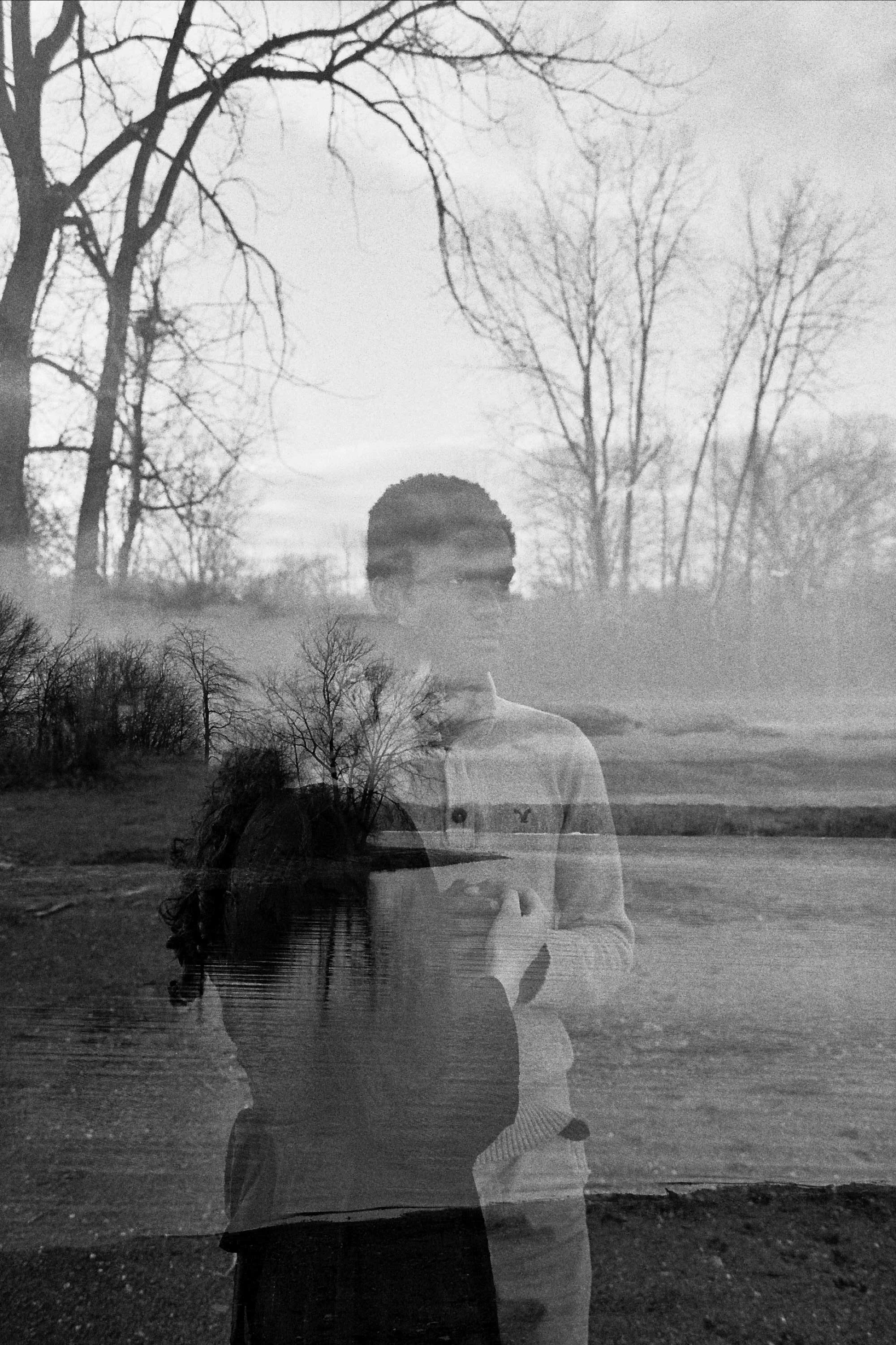 A double exposure photograph showing a young man and woman standing outdoors near a body of water, with leafless trees and a cloudy sky in the background. The image is in black and white.