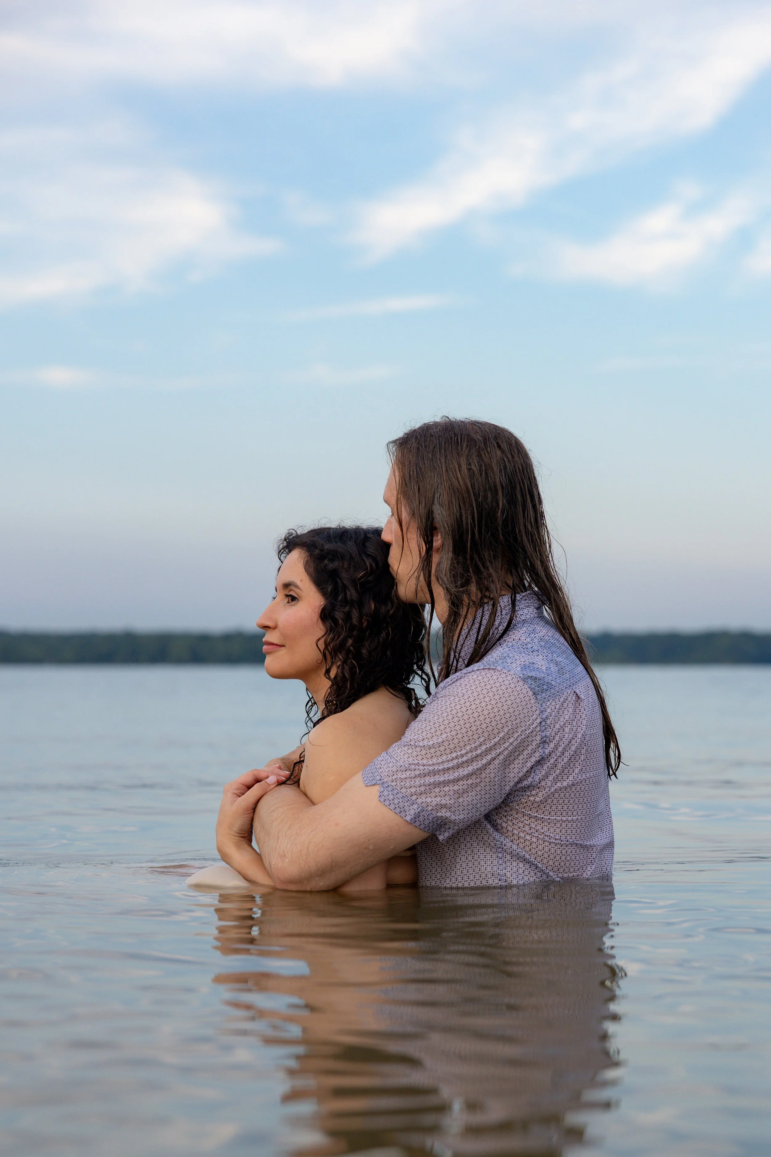 A couple is standing in a body of water, embracing each other with their arms wrapped around one another. The man is kissing the woman on her forehead, and she has a serene expression. The water reaches just below their shoulders, and the background 