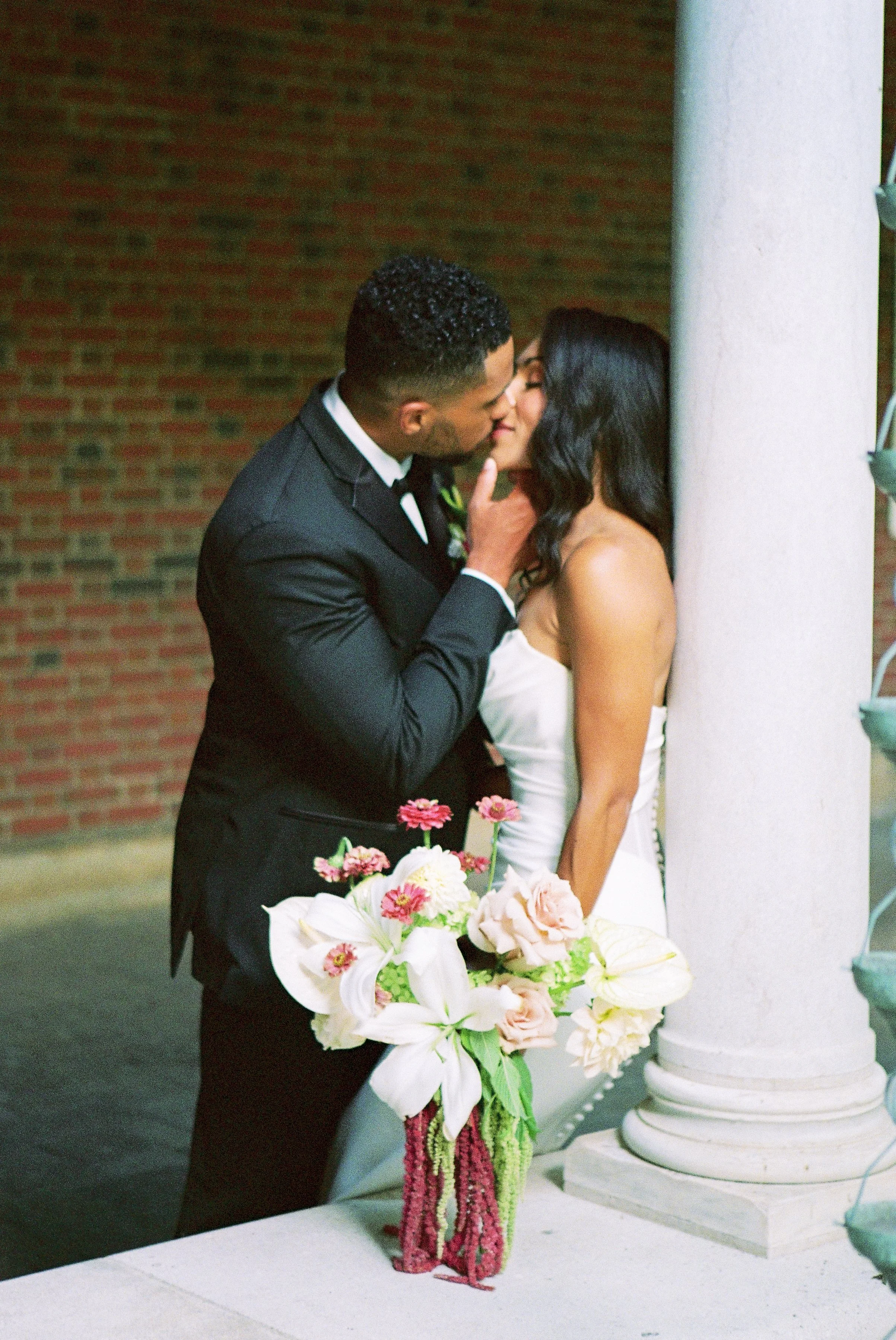 A man in a black tuxedo and a woman in a white dress sharing a kiss next to a bouquet of pink and white flowers, with a brick wall and a white column in the background.