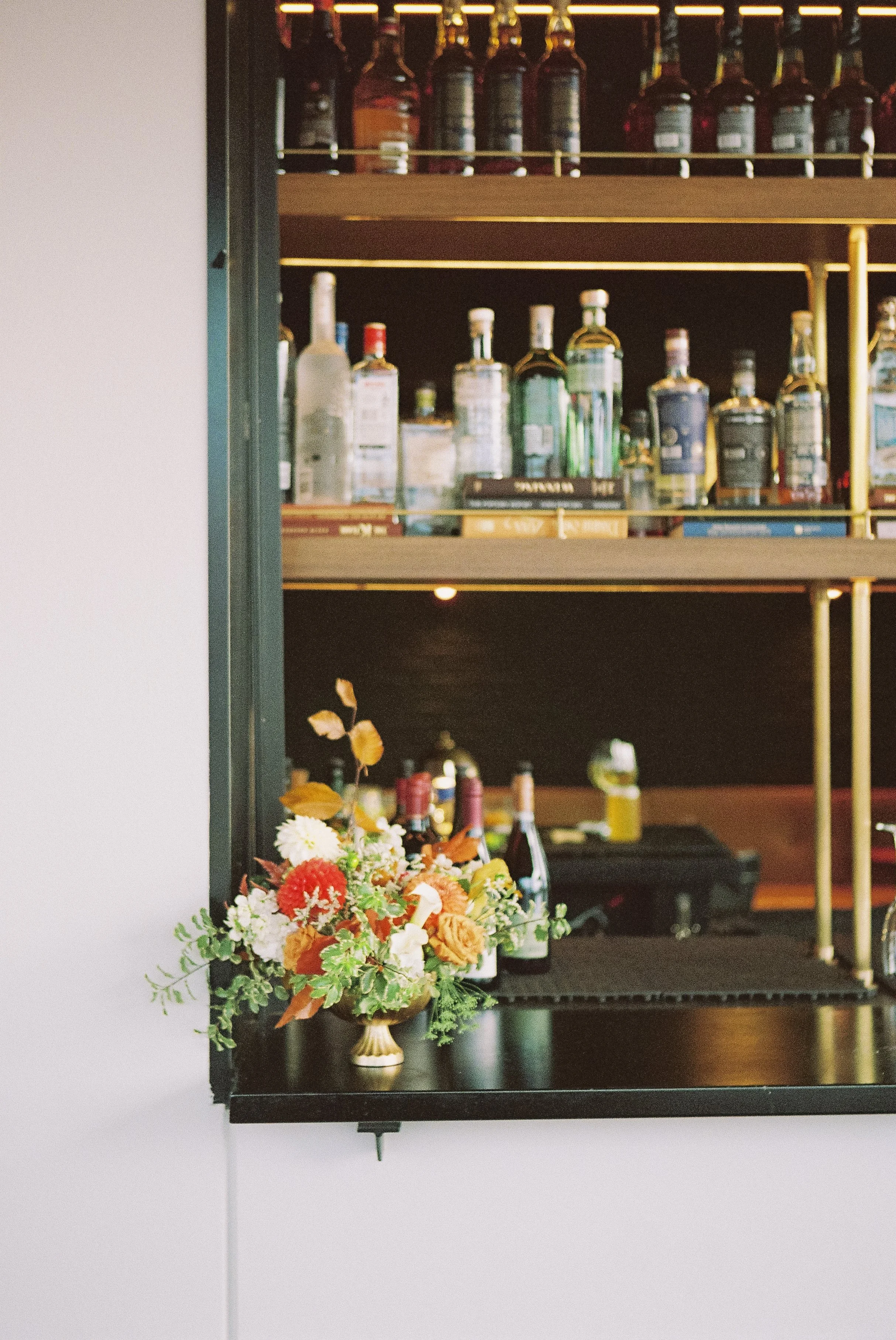 A bar shelf displaying various bottles of alcohol and a floral arrangement on the counter in front of it.
