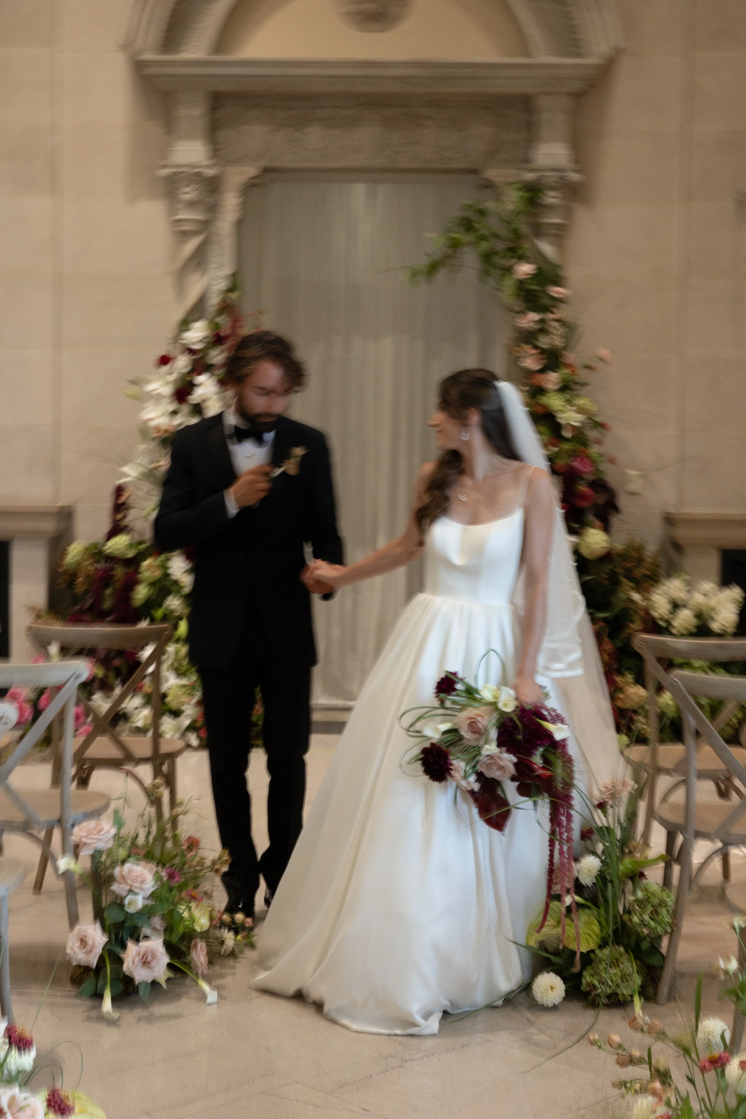 A bride and groom standing hand in hand at their wedding ceremony, surrounded by floral decorations, with the bride holding a bouquet and wearing a white wedding dress, and the groom in a black tuxedo.