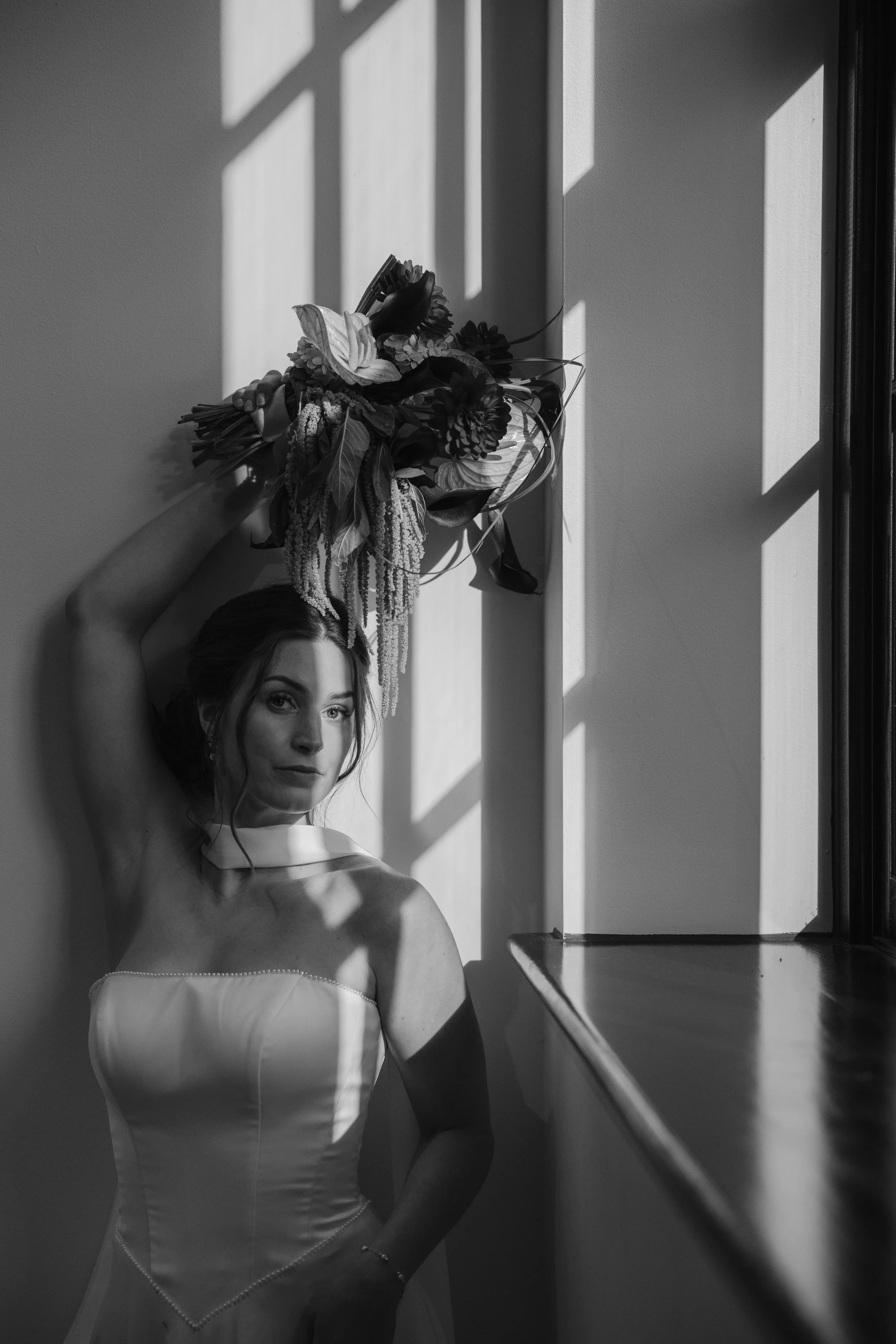 A woman in a wedding dress holding a bouquet of flowers near a window with sunlight casting shadows.
