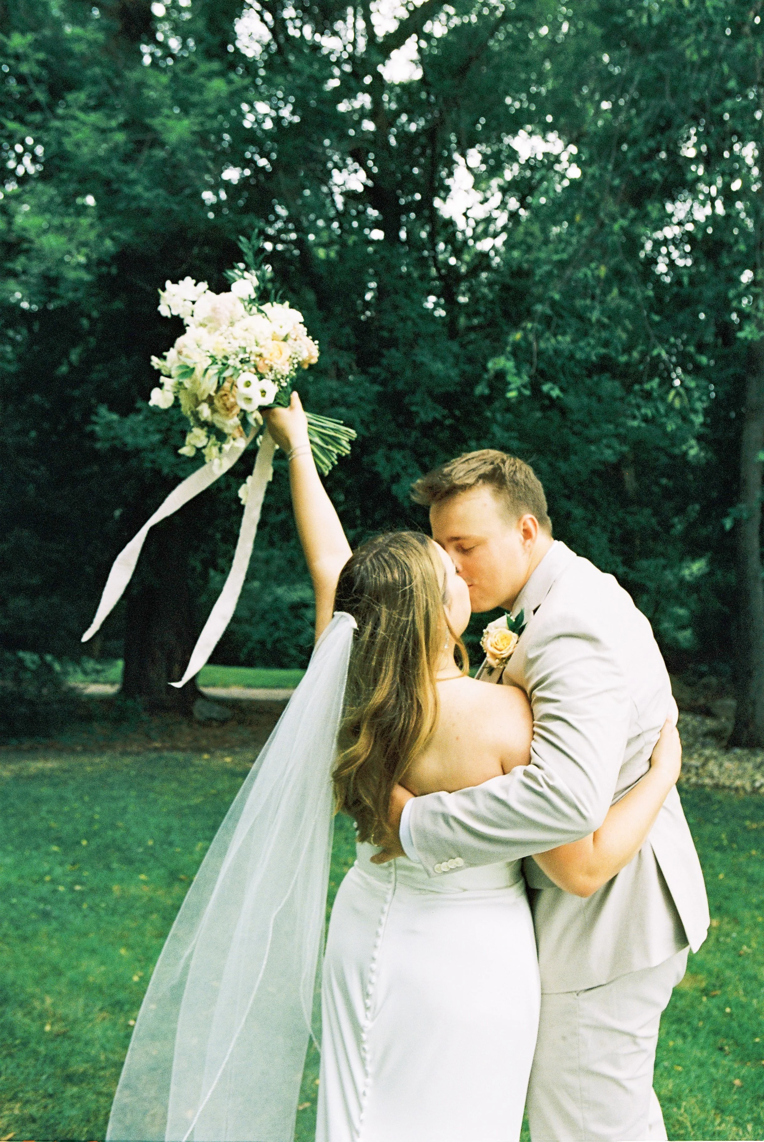 A bride and groom share a kiss outdoors, with the bride raising a bouquet of flowers in the air.