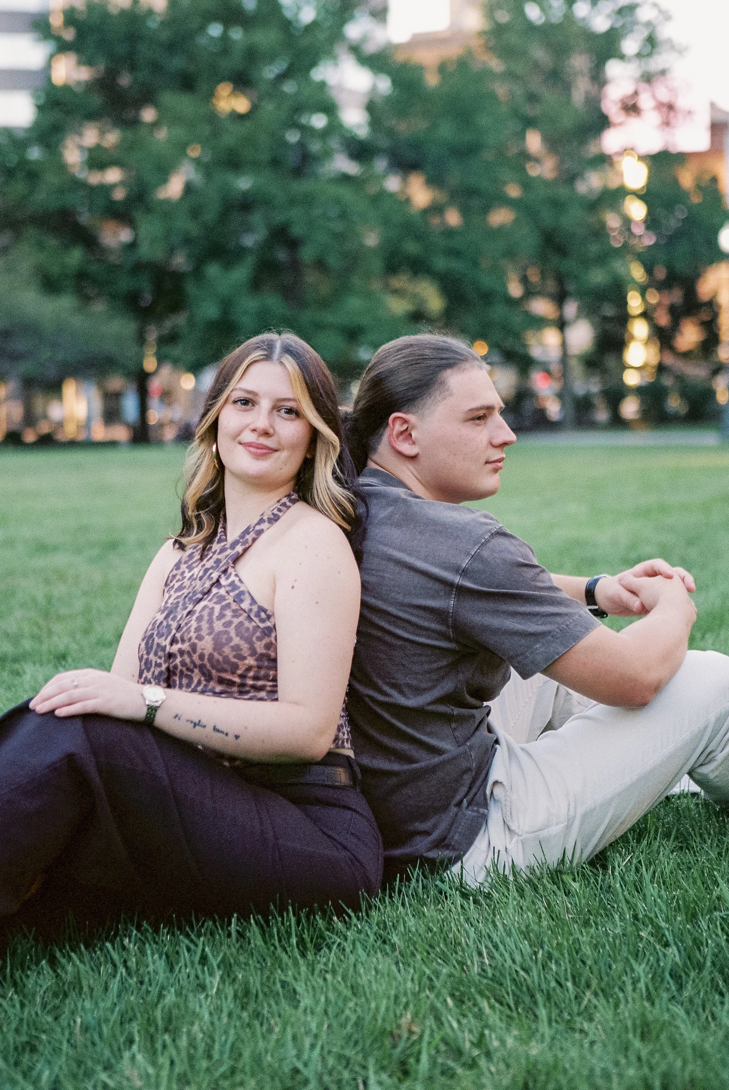 A woman and a man sitting back-to-back on a grassy field in a park, with trees and city buildings in the background during sunset.