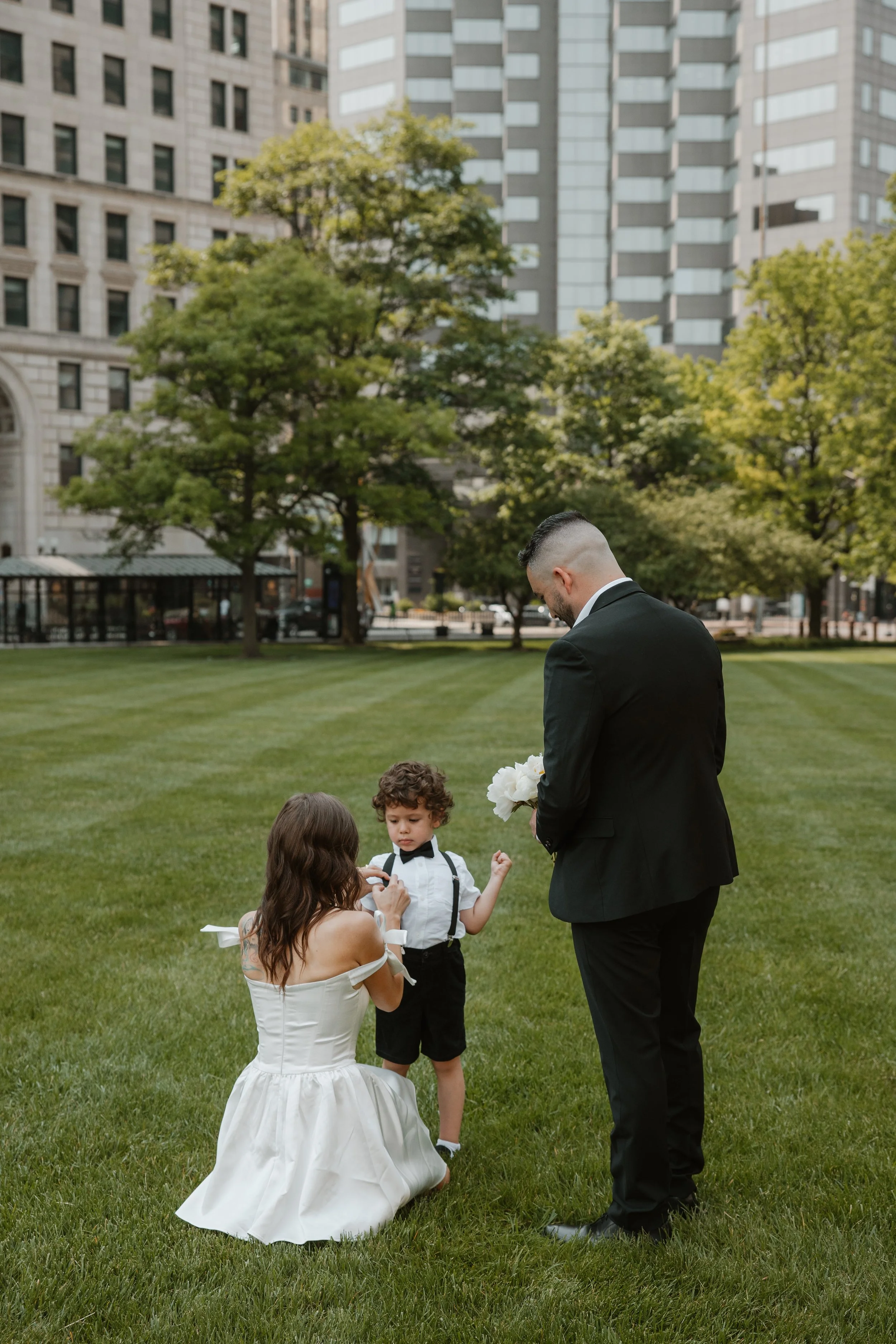 A bride, groom, and young boy in a city park with green grass and trees, during a wedding ceremony.