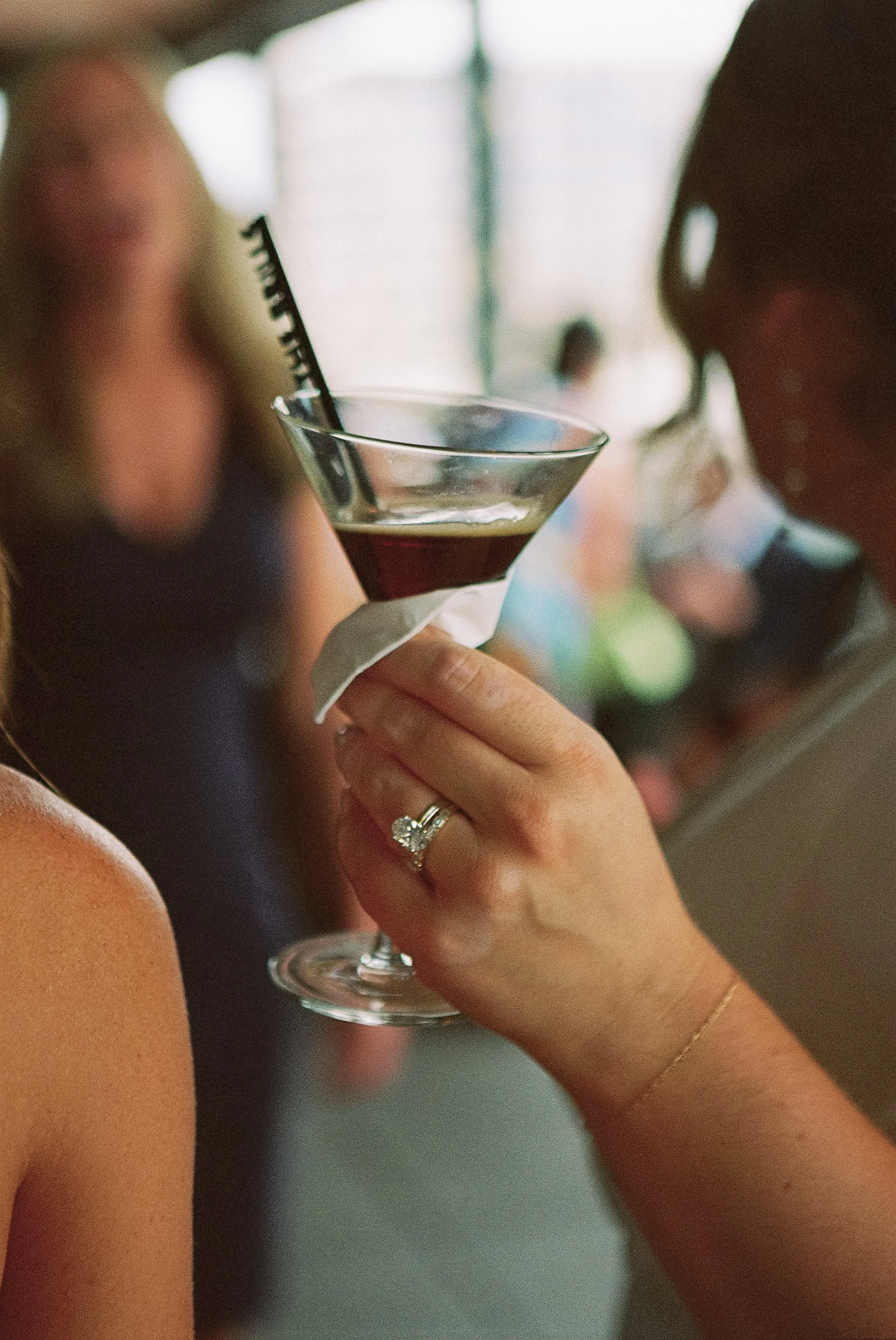 A woman wearing a ring is holding a martini glass with a dark red cocktail, garnished with a black straw, at a social event. In the background, there are blurred figures of people.