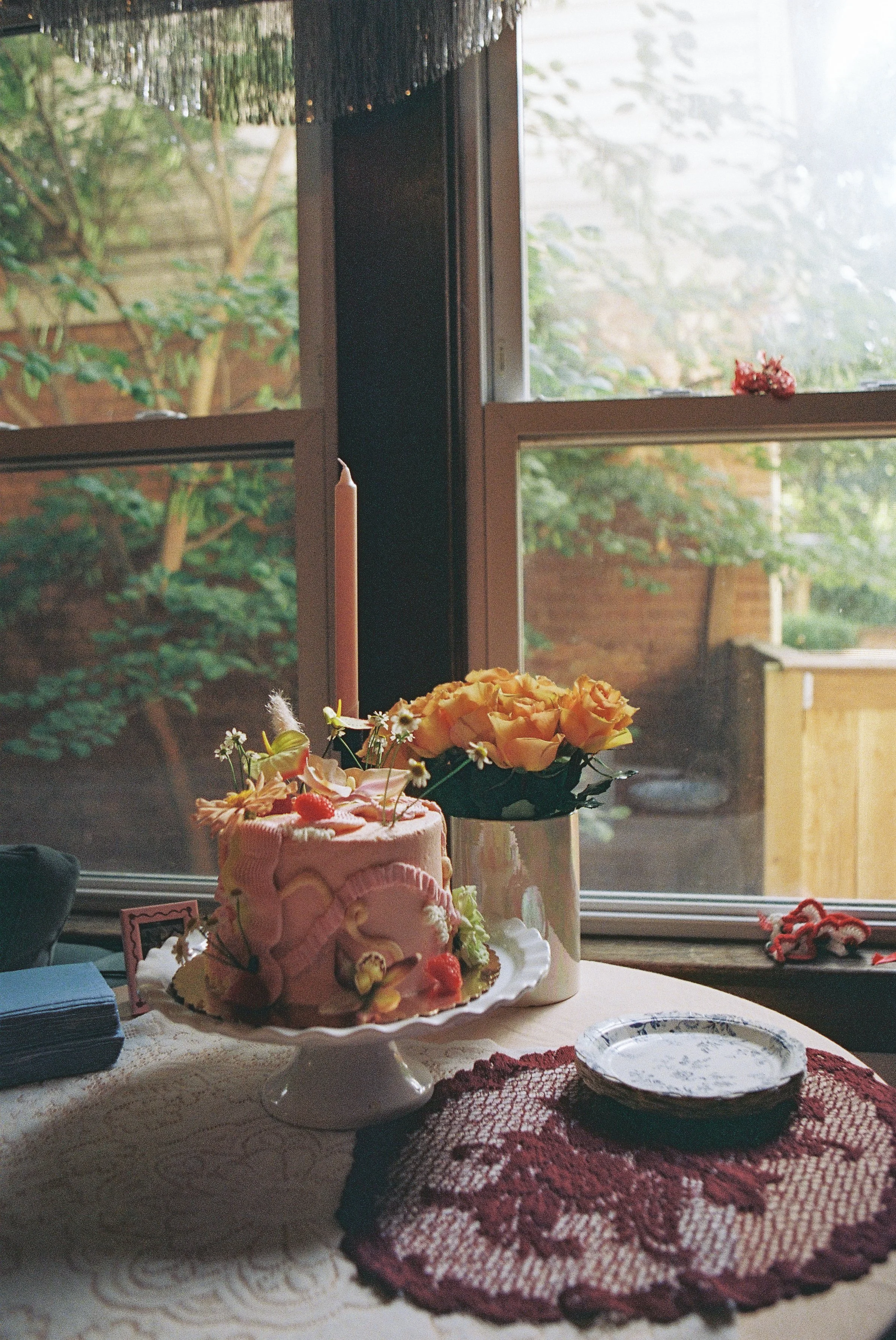 A decorated birthday cake on a cake stand with pink icing, strawberries, and flowers, next to a vase of yellow roses on a table near a window with outdoor greenery.
