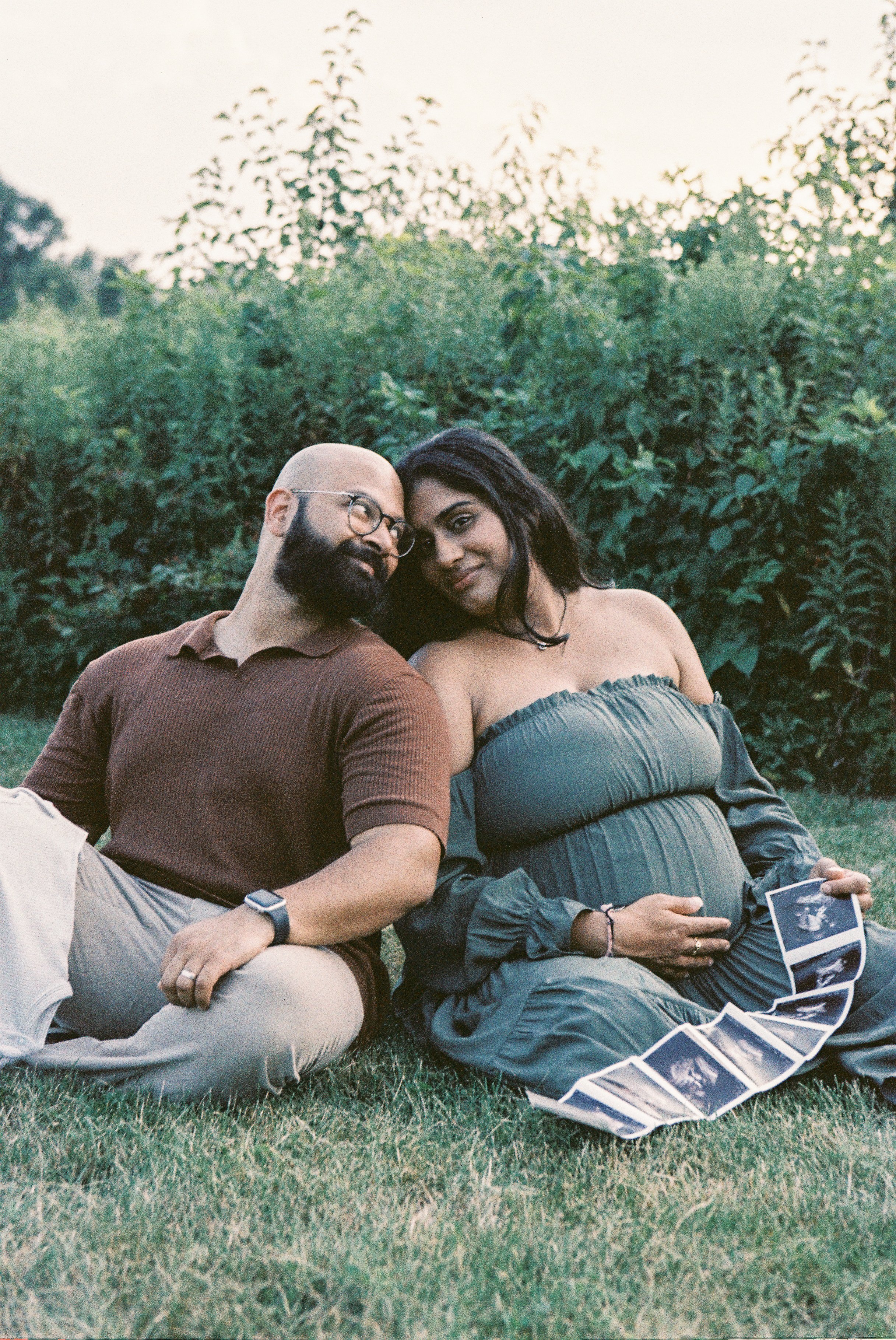 A couple sitting on the grass during sunset, with a pregnant woman holding ultrasound photos, and a background of greenery.