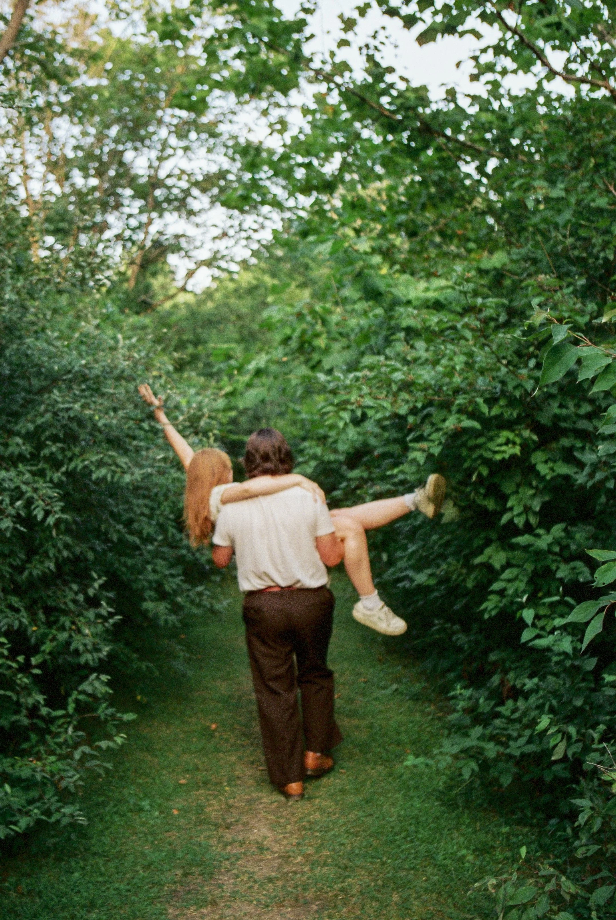 A person walking down a narrow green pathway surrounded by dense green bushes and trees while carrying a woman in his arms, with her arms and legs outstretched and one arm raised.