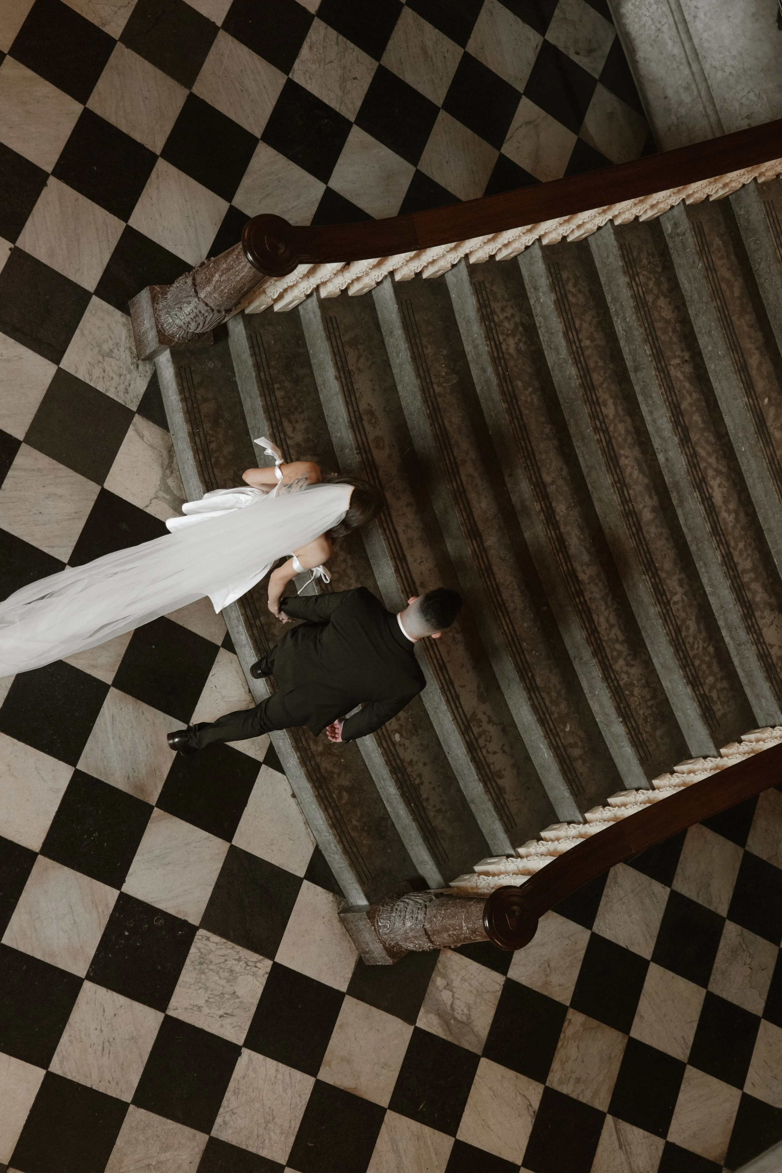 A bride in a white wedding dress and a groom in a black suit walking up a staircase with a checkered black and white tile floor surrounding them.