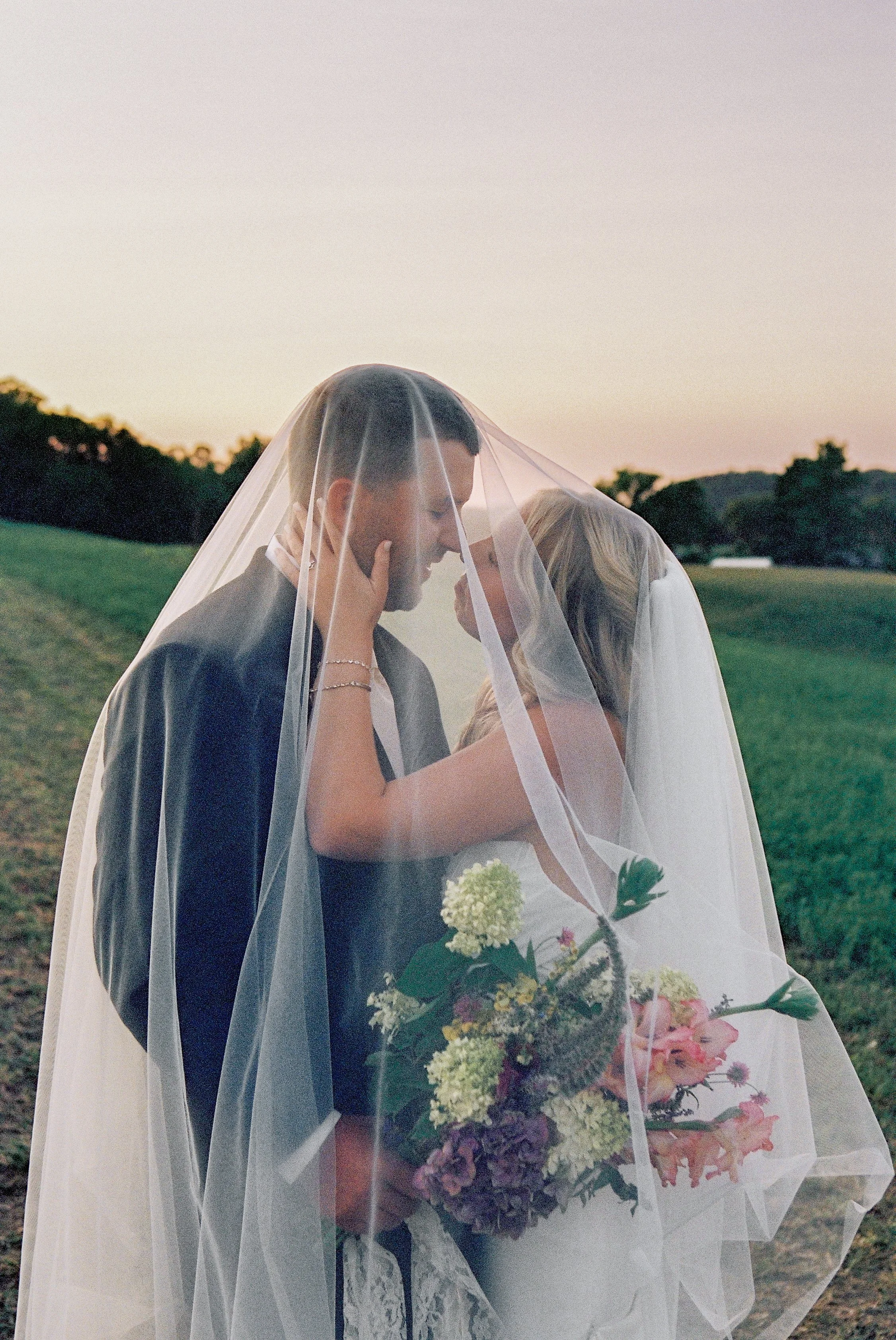 A bride and groom sharing a kiss under a wedding veil, standing outdoors on a grassy field at sunset, with the bride holding a bouquet of flowers.