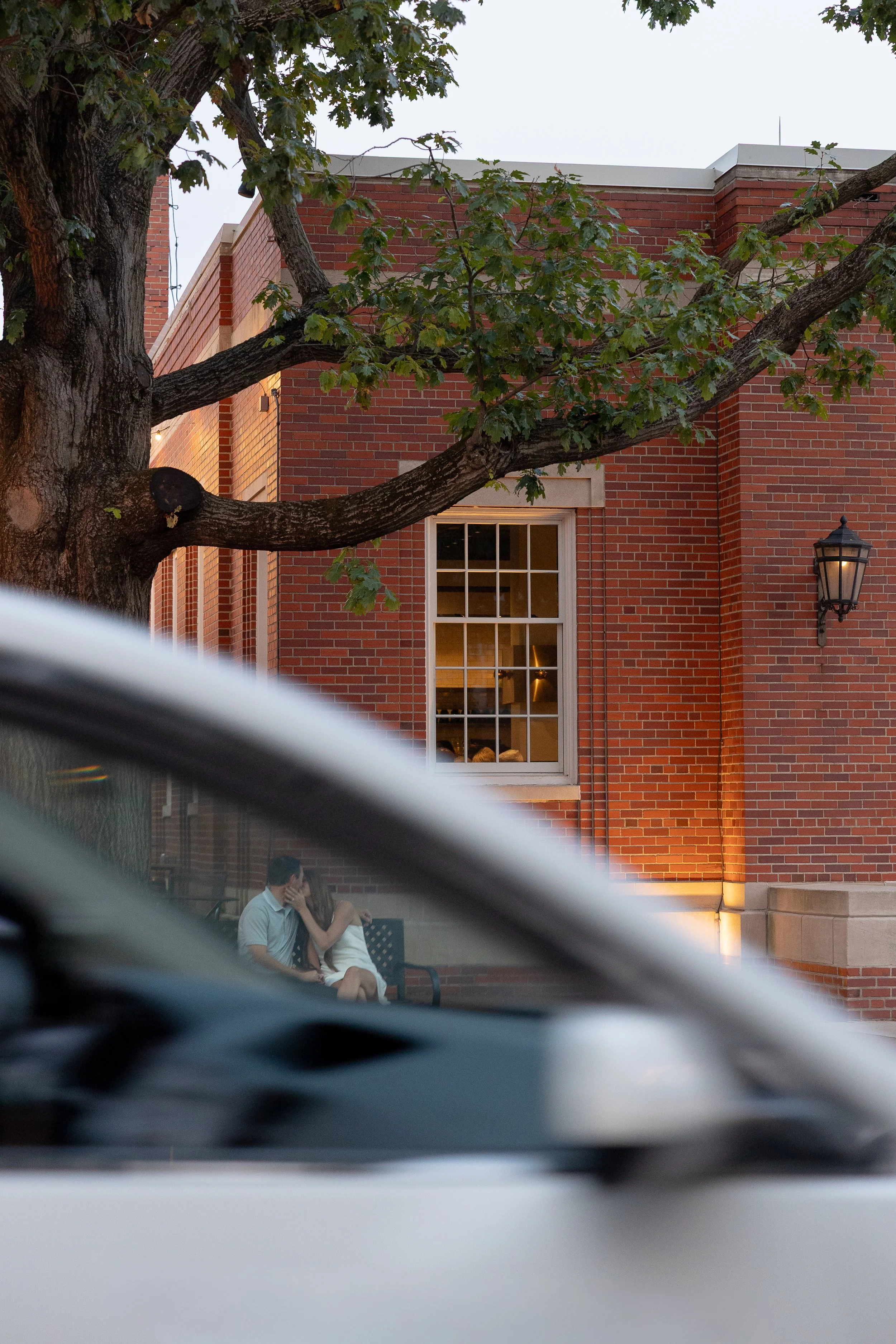 A couple sitting on a bench outside a brick building, with a large tree and window visible in the background. The photo is taken from behind a parked car, with part of the car in the foreground.