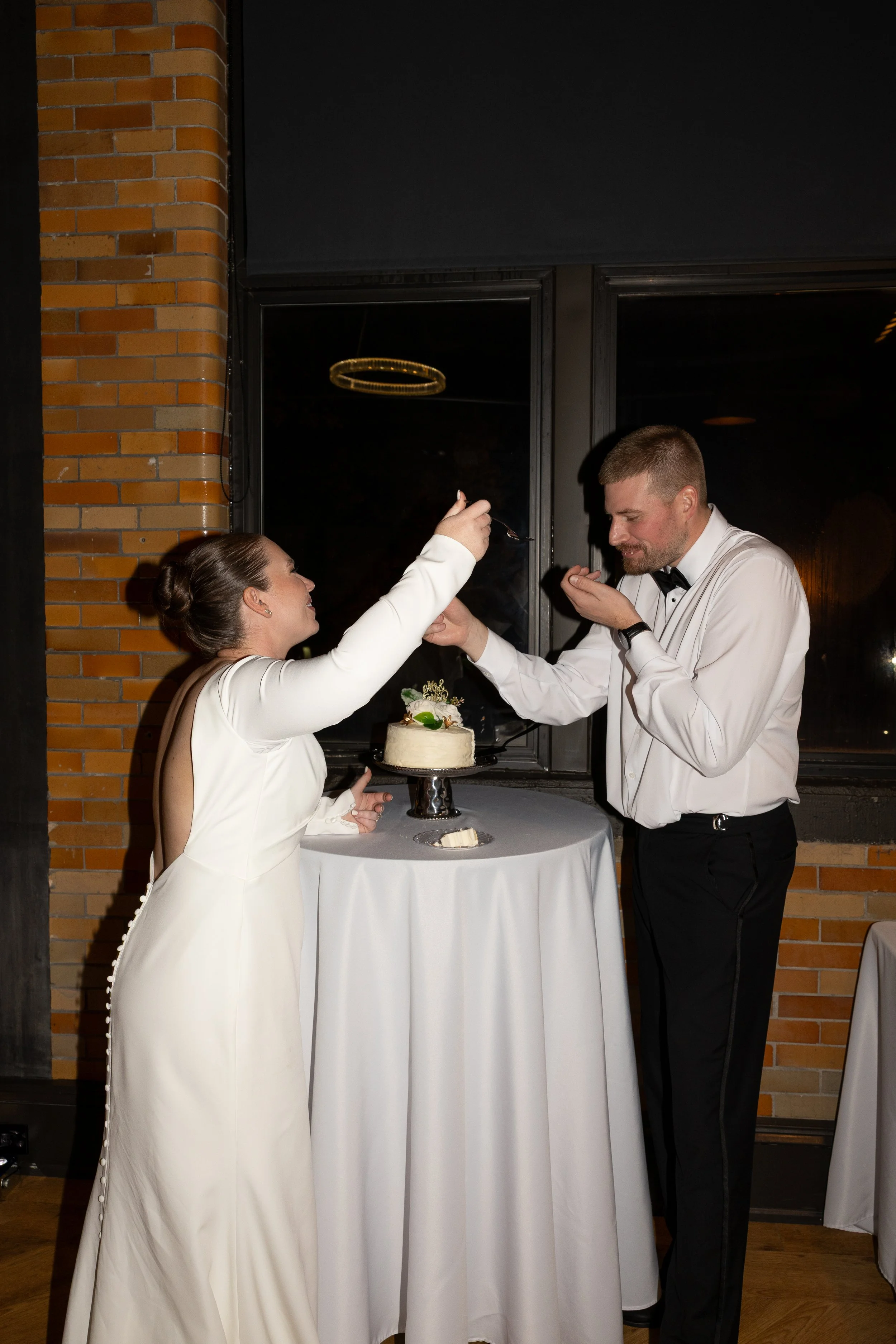 A bride and groom sharing a piece of wedding cake at a reception, with the bride feeding cake to the groom, standing at a table with a white tablecloth and a small wedding cake.