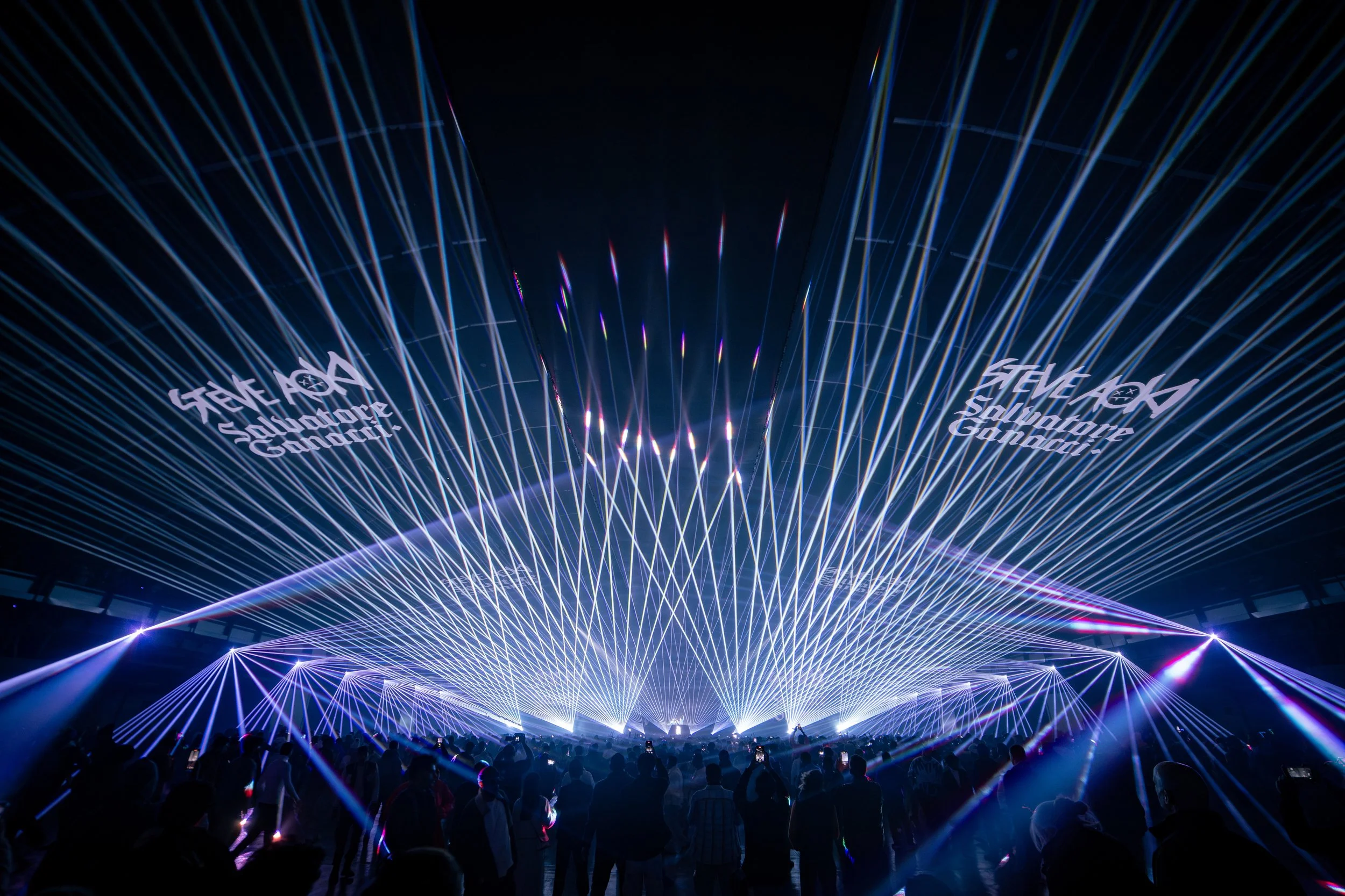 A large crowd watching a colorful laser light show with bright blue and purple beams and the text 'Steve Aoki Salvador Gunduz' projected in the sky.