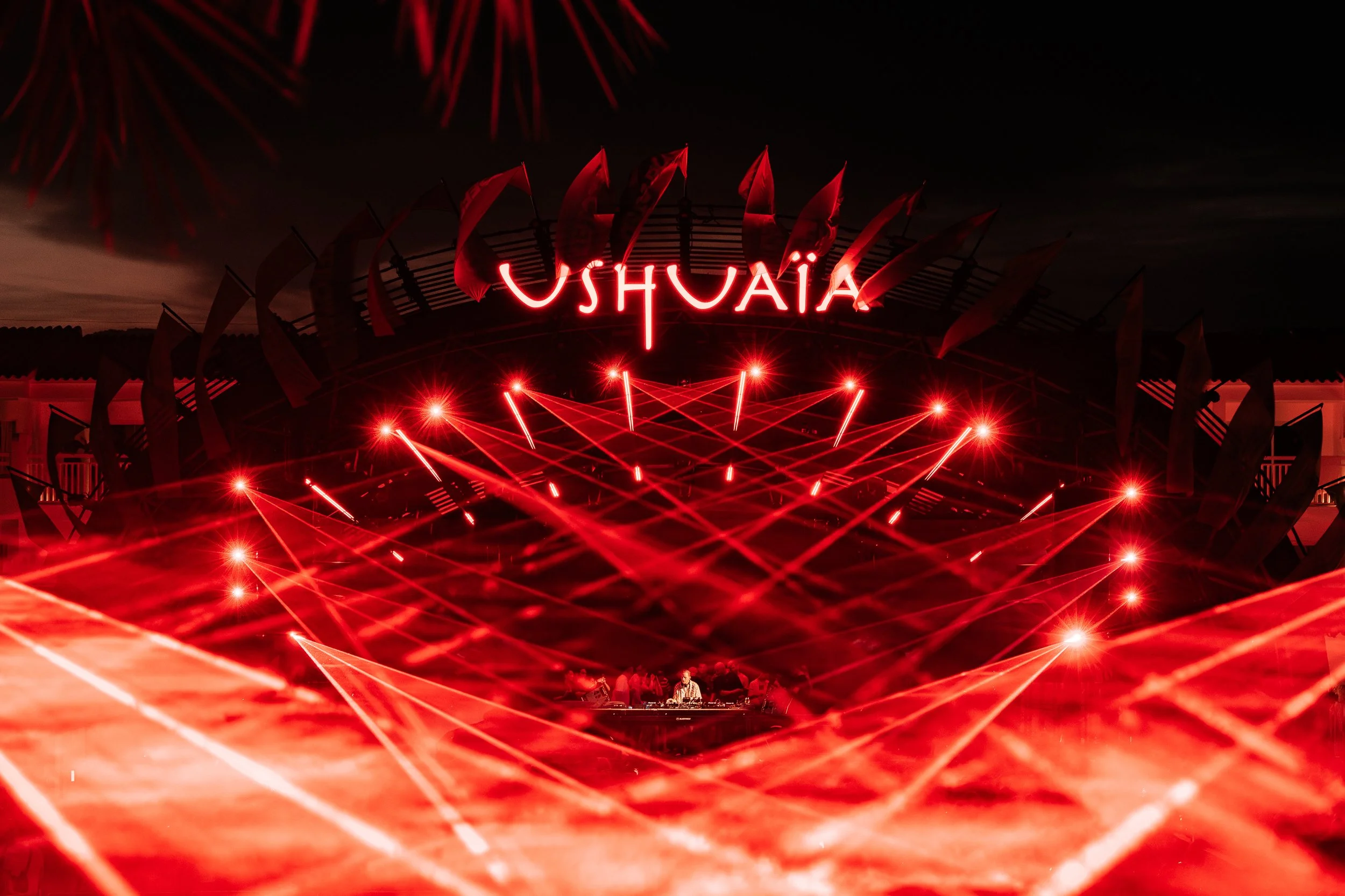 Red fireworks in the night sky above a stage with bright red laser lights and the word 'USHUAÏA' neon sign, with people visible at the bottom.