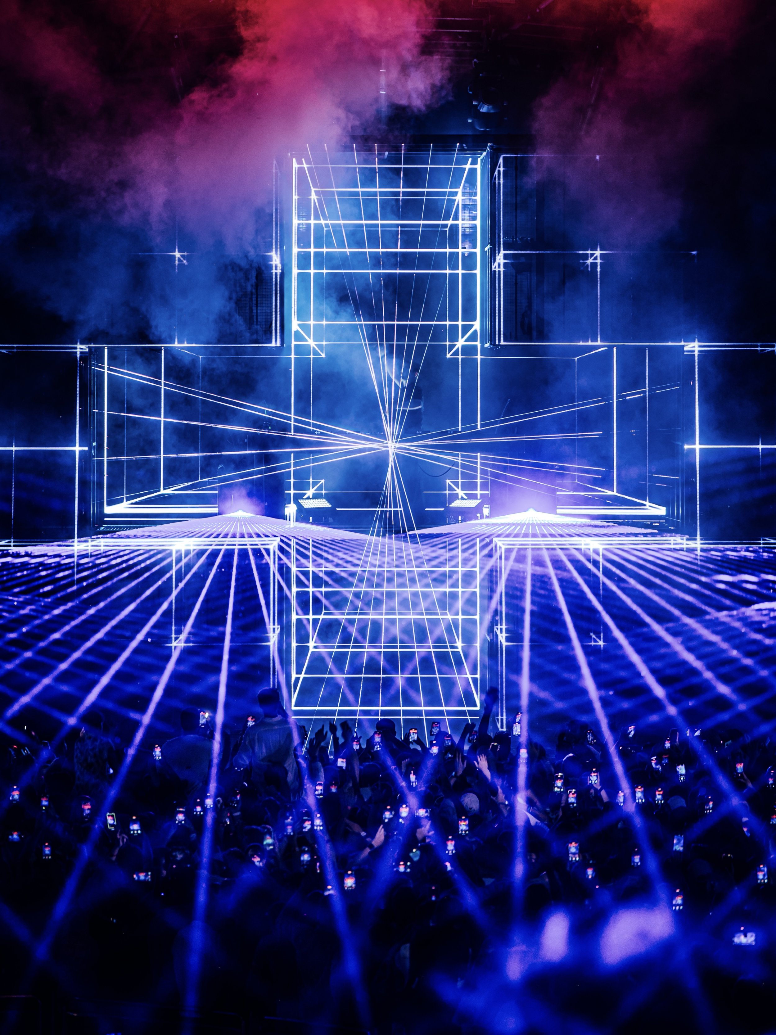A crowd of people at a concert or event, all holding up their phones to take pictures or record, with a large, illuminated geometric light installation on stage. The stage features a three-dimensional wireframe design with blue and purple lighting, e