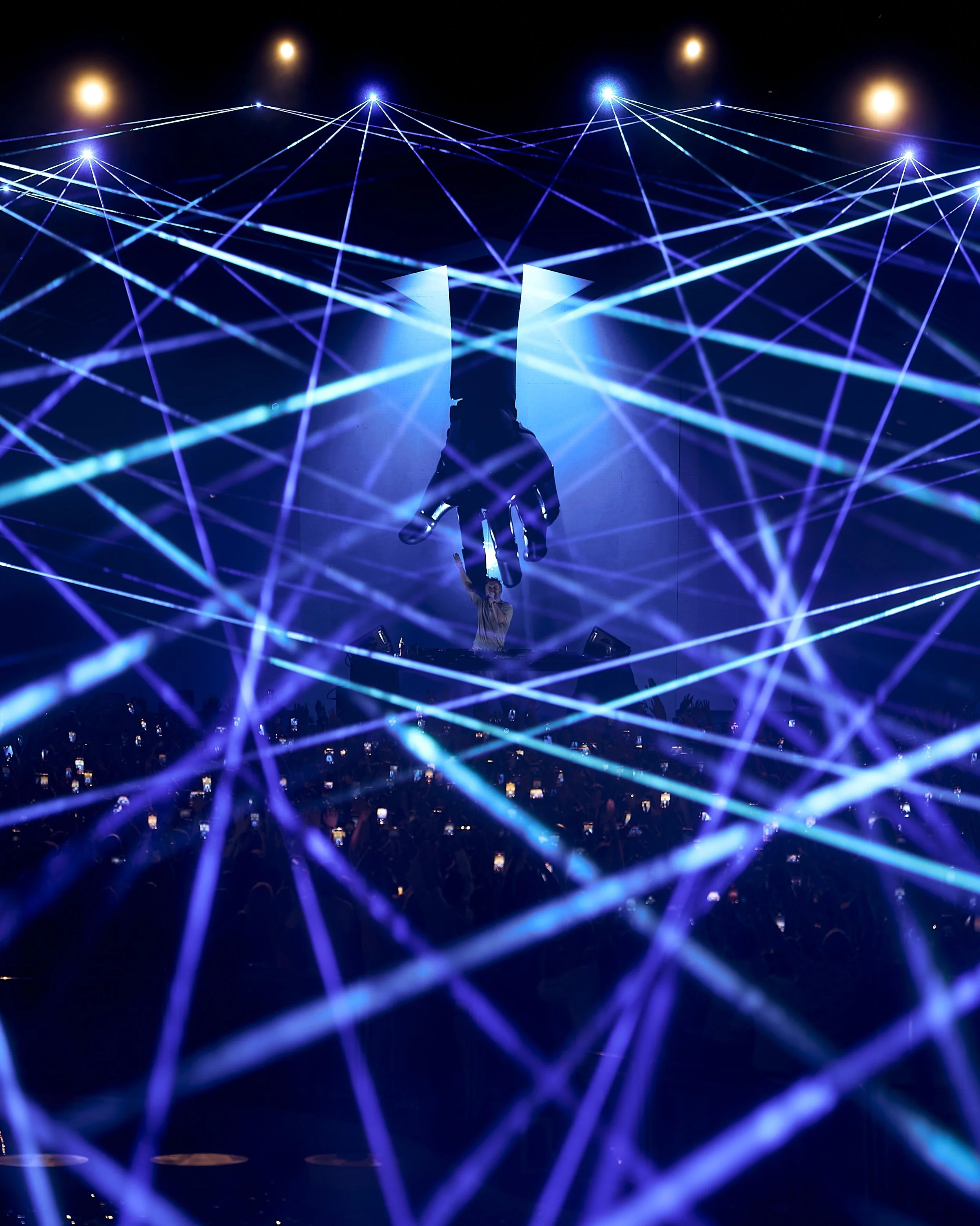 Performance stage with a DJ, colorful laser lights forming a web-like pattern, and a large floating hand atop the DJ, with audience members capturing the moment on their phones.