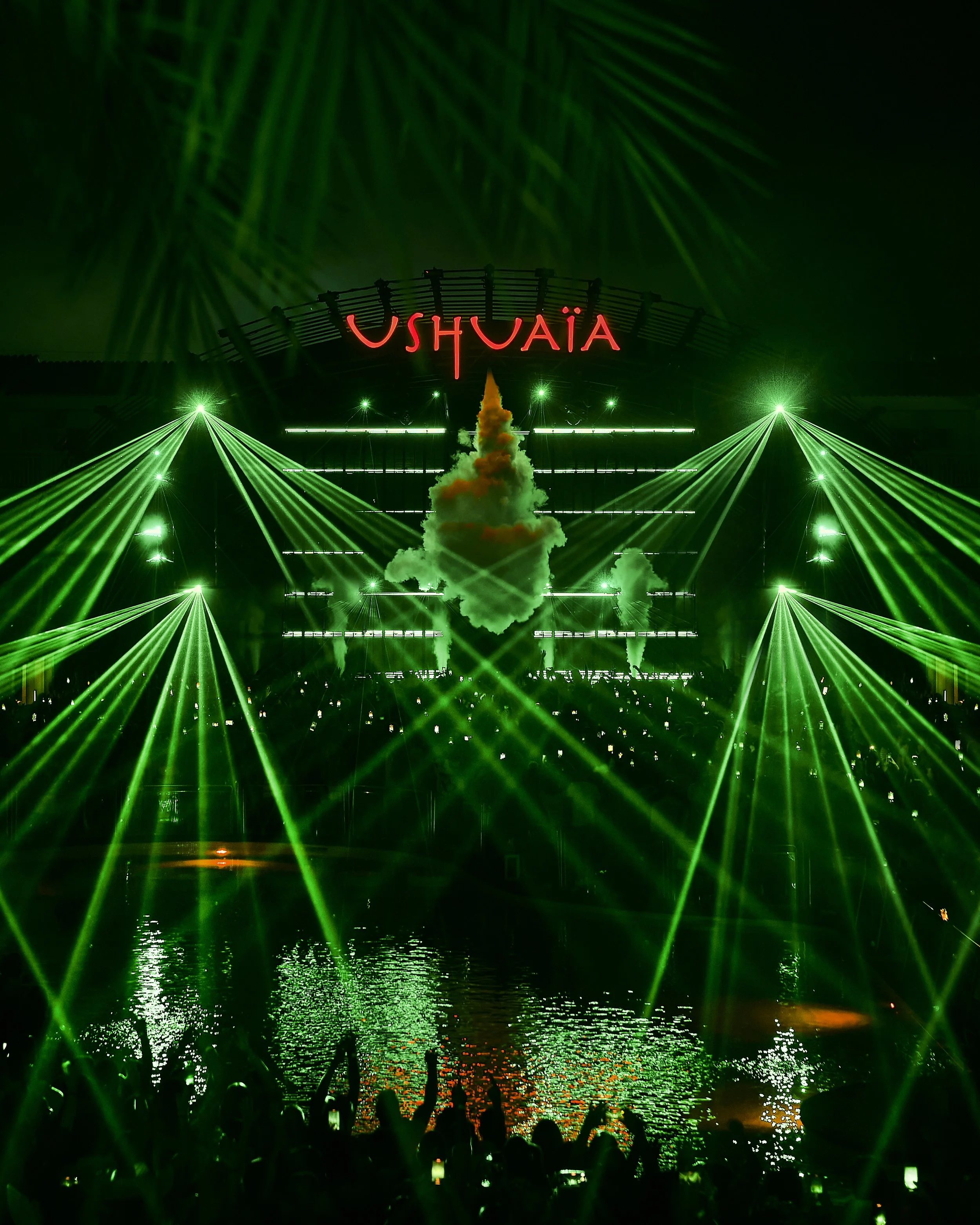 Nighttime scene of a laser light show over water with green laser beams and smoke. A large red neon sign reads 'USHAIA' above the water, with silhouettes of audience members holding candles or lights in the foreground.